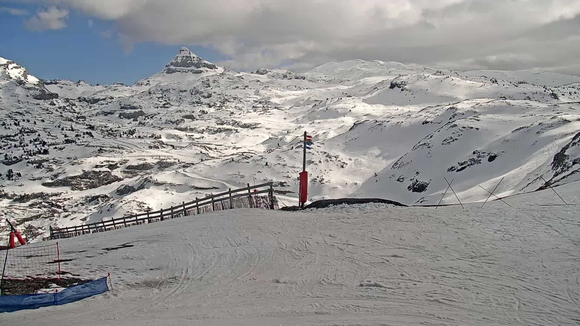 A snowy mountain range is visible under a partly cloudy sky, with a ski slope in the foreground featuring a wooden fence and safety markers.
