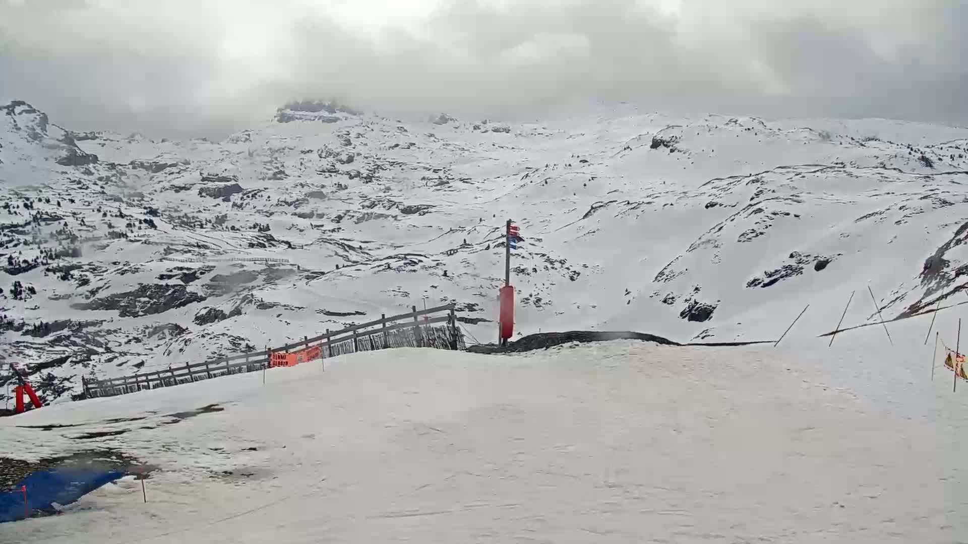 A snow-covered mountain landscape under an overcast sky, with a small section of a ski slope in the foreground.