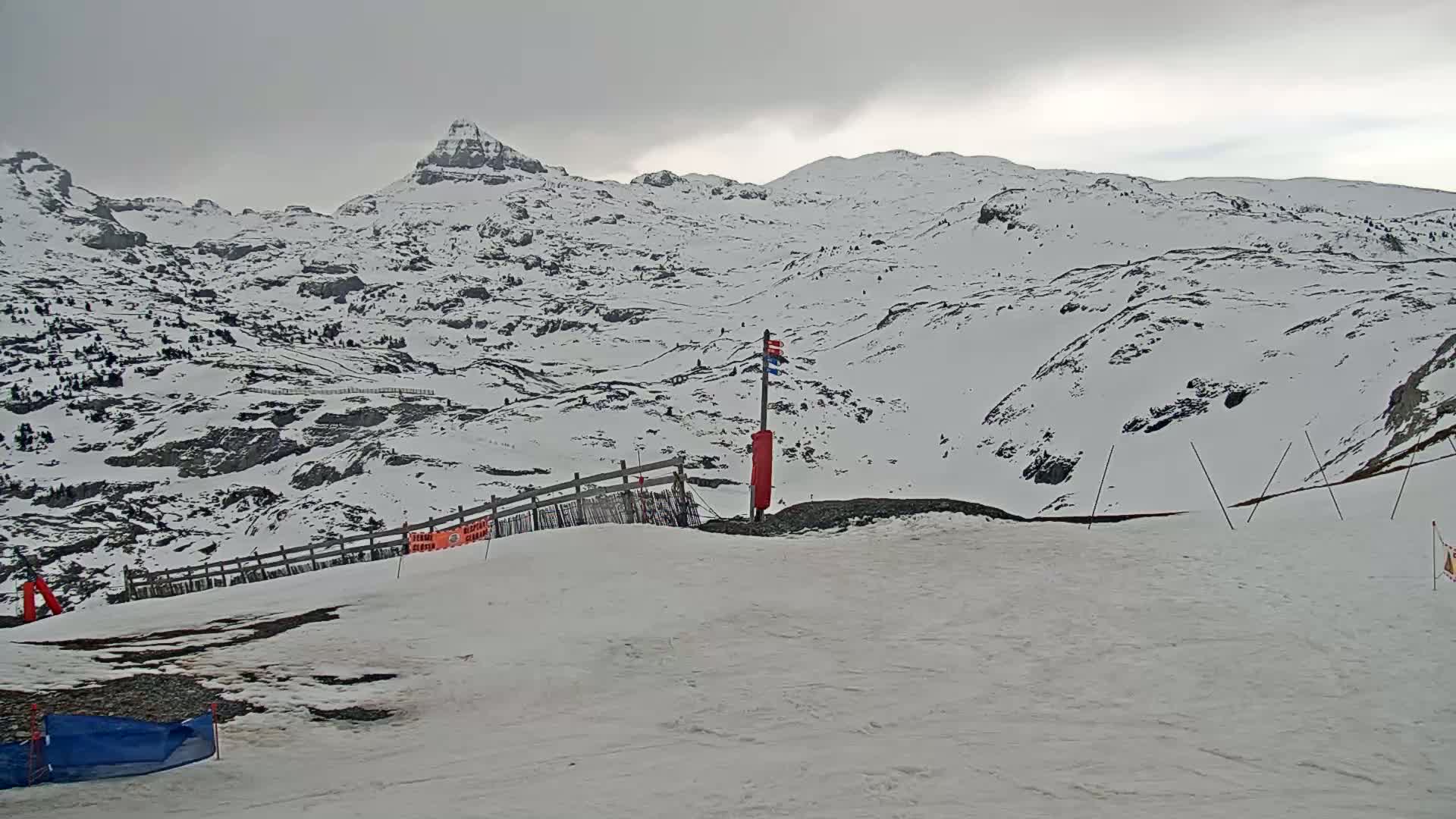 A snow-covered mountain range is visible under an overcast sky, with a section of a ski slope in the foreground.