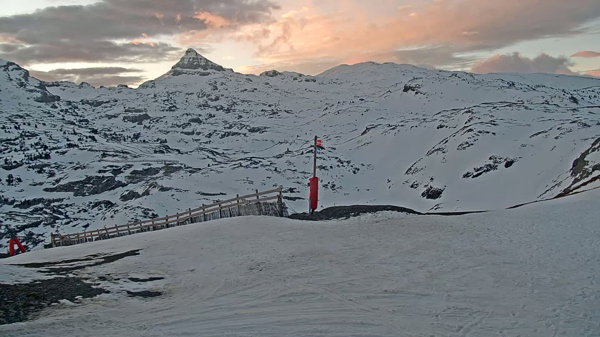 A snow-covered mountain range at sunset, with a wooden fence and a red marker visible in the foreground.