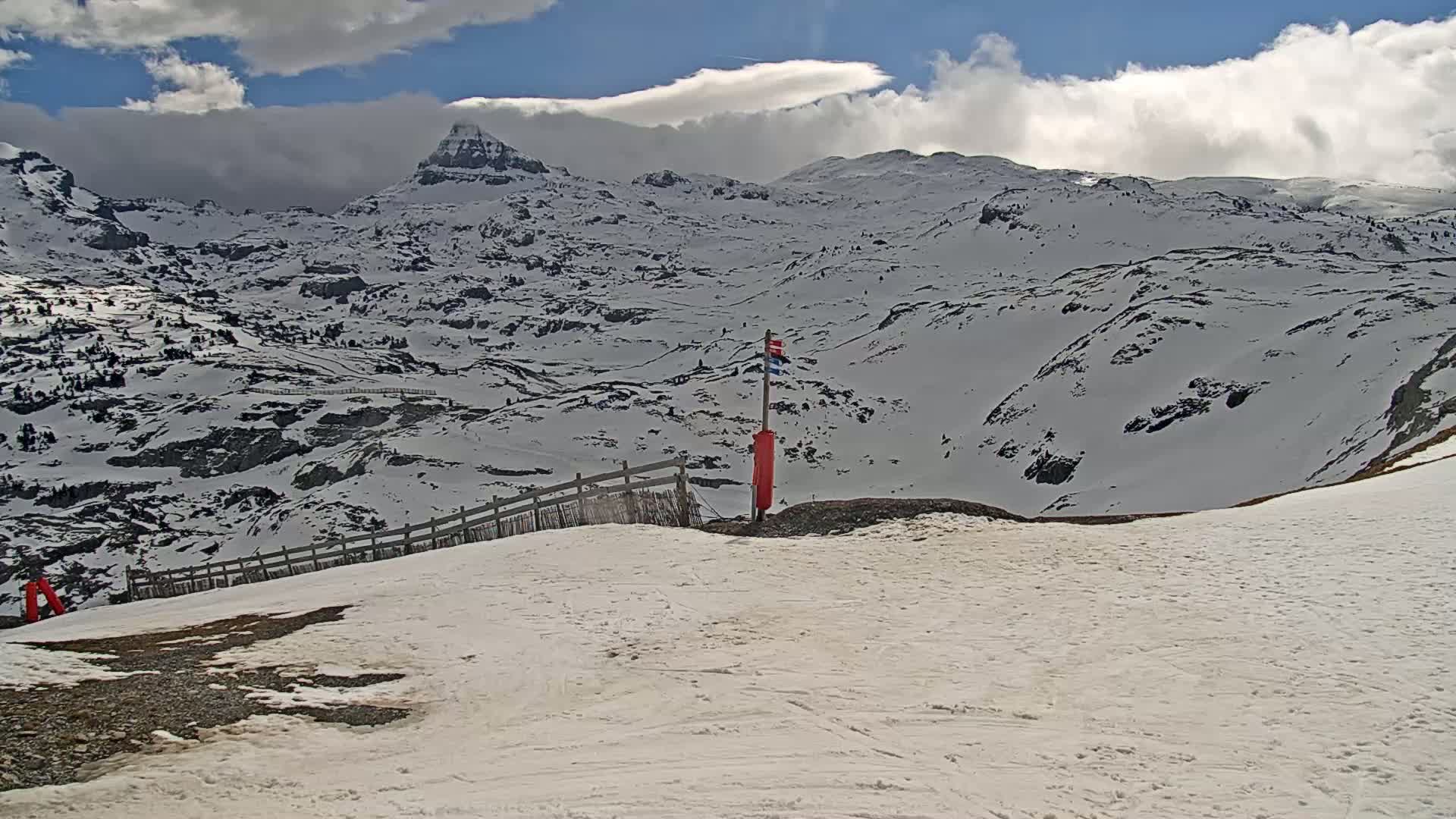 A snow-covered mountainous landscape under a partly cloudy sky, with a short wooden fence and a red marker visible in the foreground.