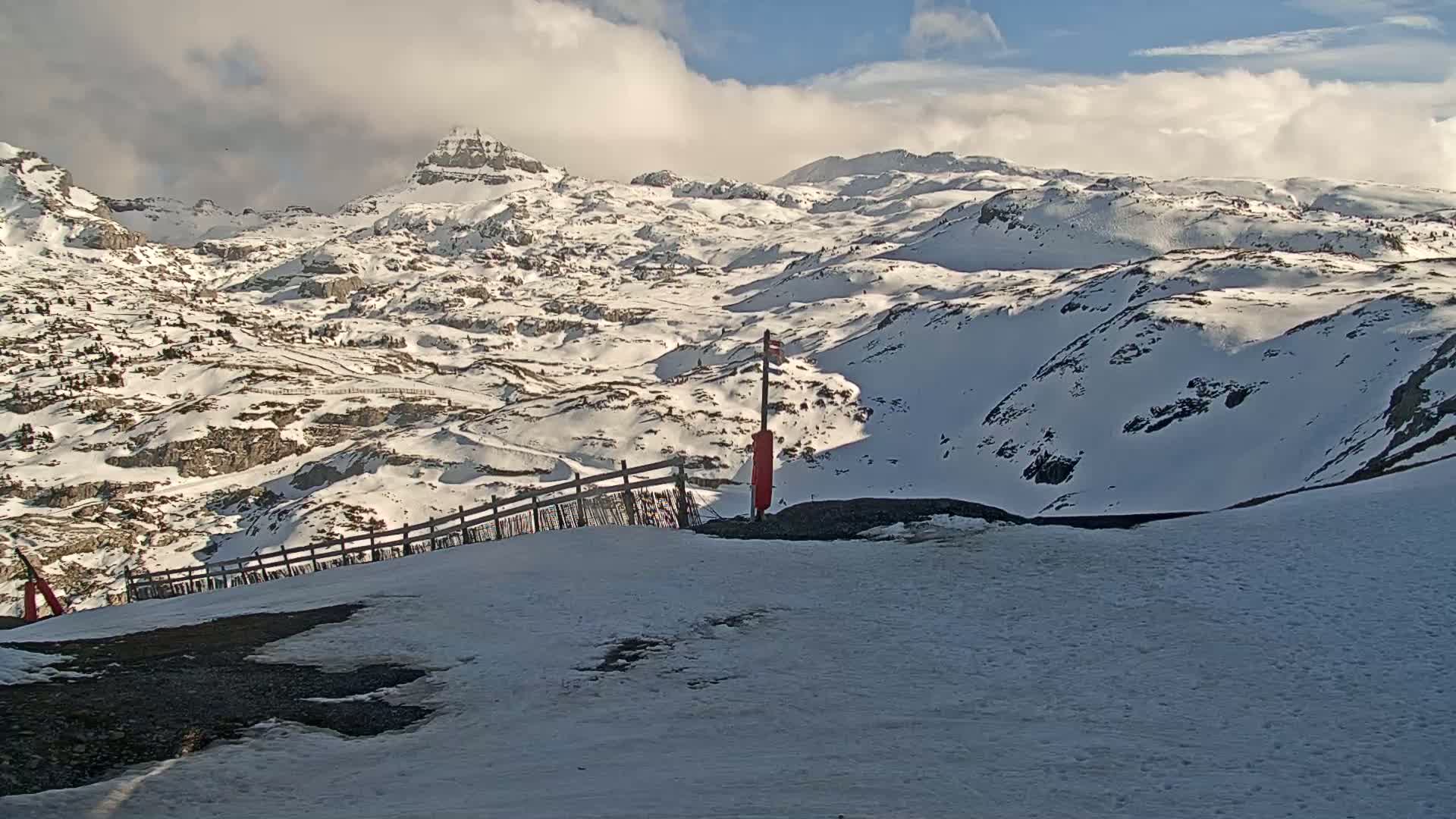 A snow-covered mountain range under a partly cloudy sky is seen from a snowy vantage point with a short wooden fence.