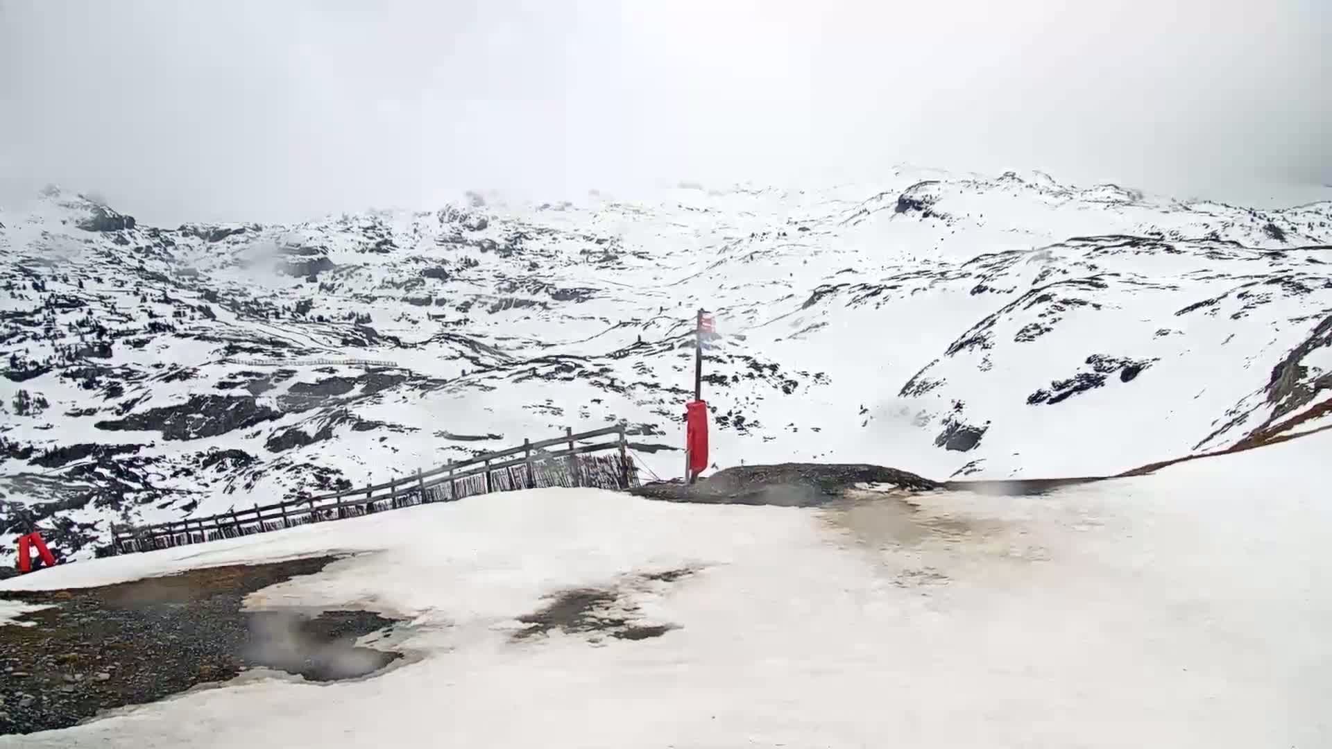 A snow-covered mountain range is viewed from a snowy slope on an overcast day.