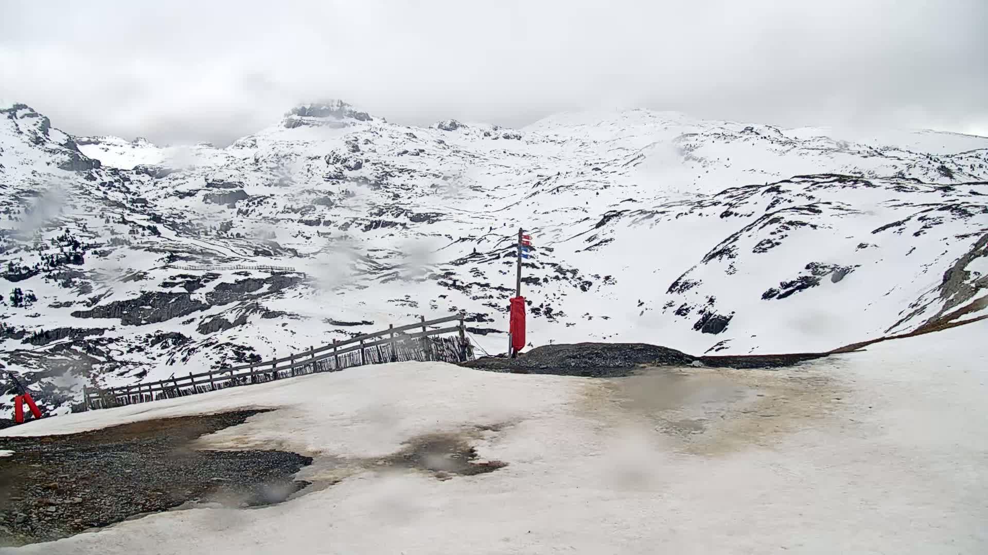 A snow-covered mountain range is visible under an overcast sky, with a small section of dark ground and a red marker visible in the foreground.