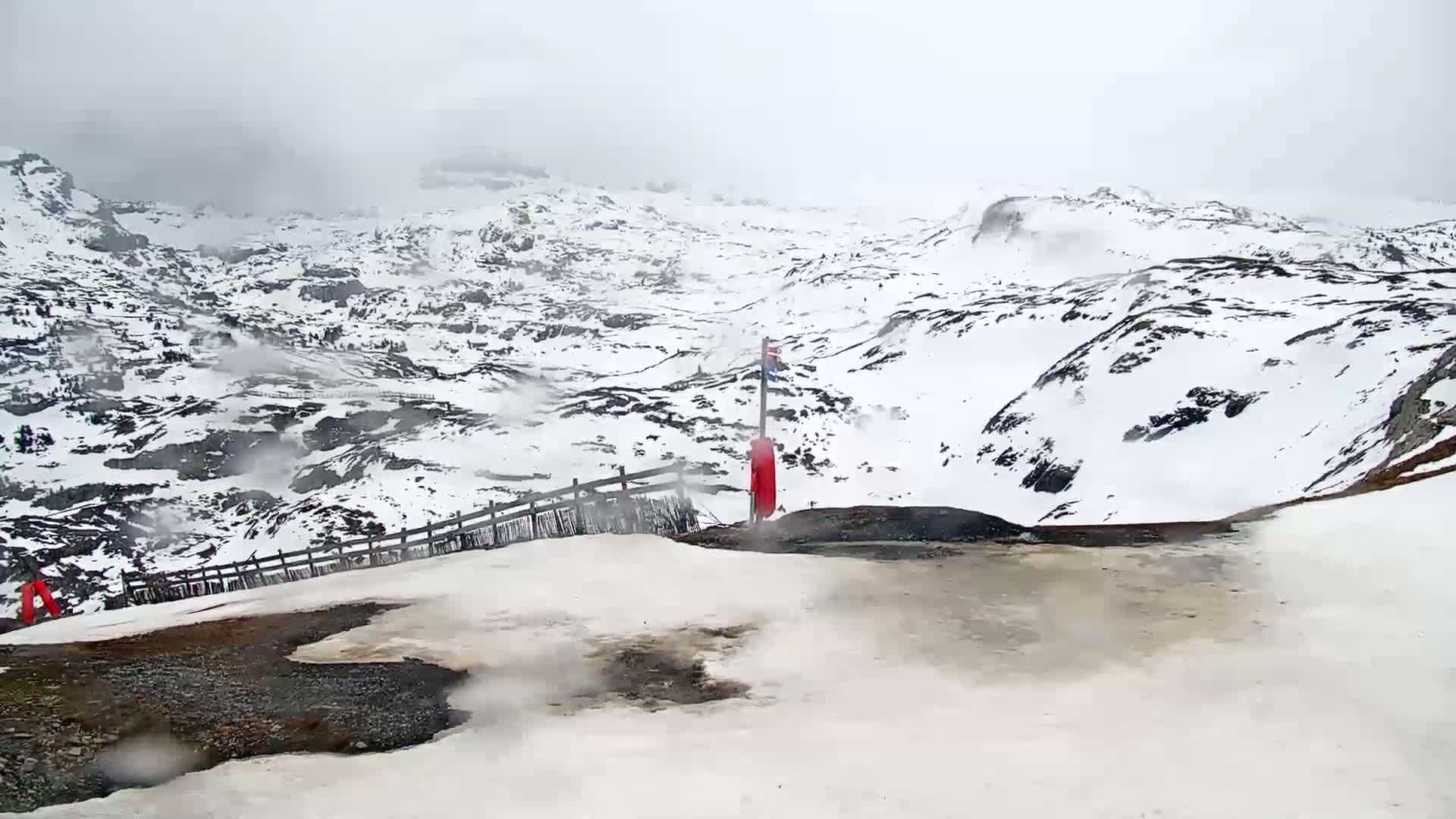 A snowy, overcast mountain range is viewed from a high vantage point, showing a wooden fence and a red object on a pole near a patch of exposed ground.