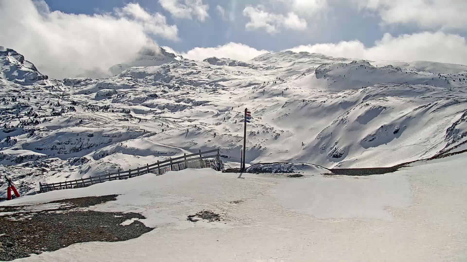 A snow-covered mountain range is visible under a partly cloudy sky, with a wooden fence and a flagpole in the foreground.