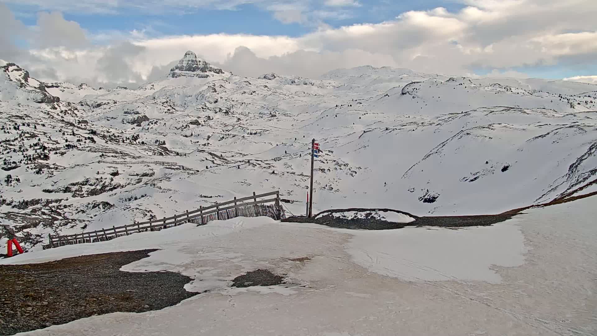 A snow-covered mountainous landscape under a partly cloudy sky, featuring a wooden fence and a flagpole near a snow-covered slope.