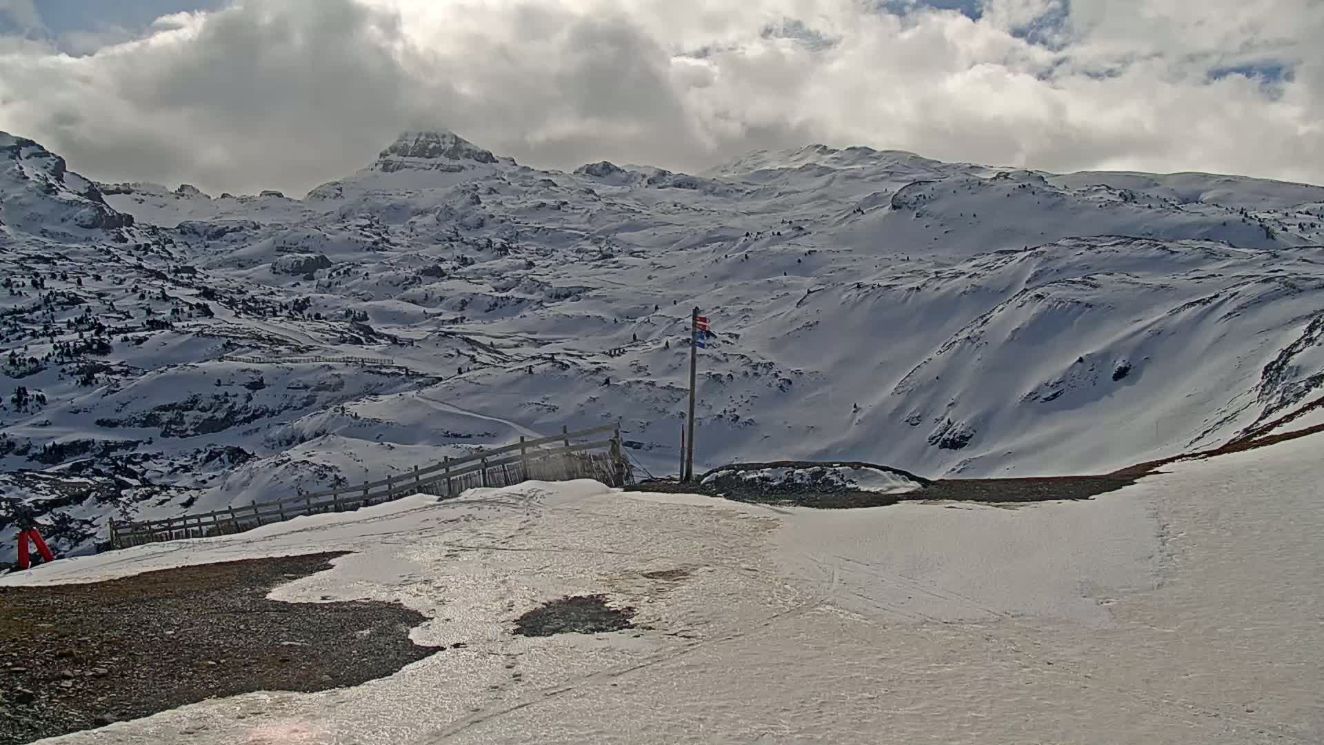 A snow-covered mountainous landscape under partly cloudy skies shows a wooden fence and a flagpole near a patch of bare ground.