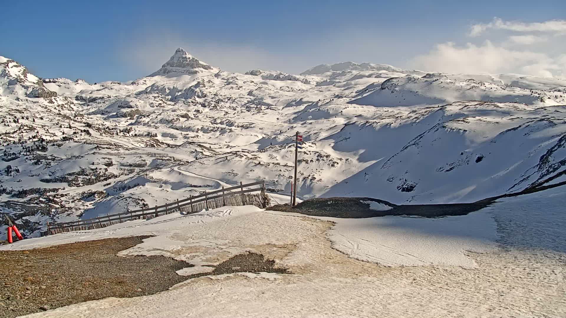 A snow-covered mountain range under a mostly clear blue sky, with a wooden fence and a marker post in the foreground.