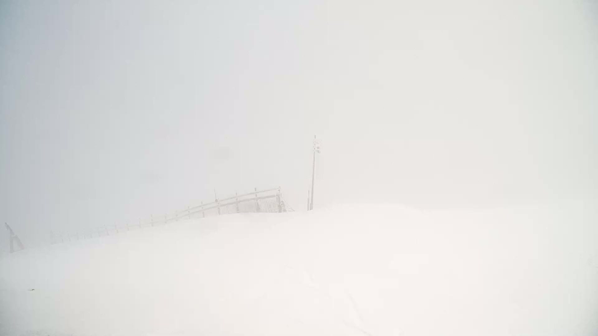 A snow-covered landscape with a partially visible wooden fence and a tall pole is shrouded in thick fog.