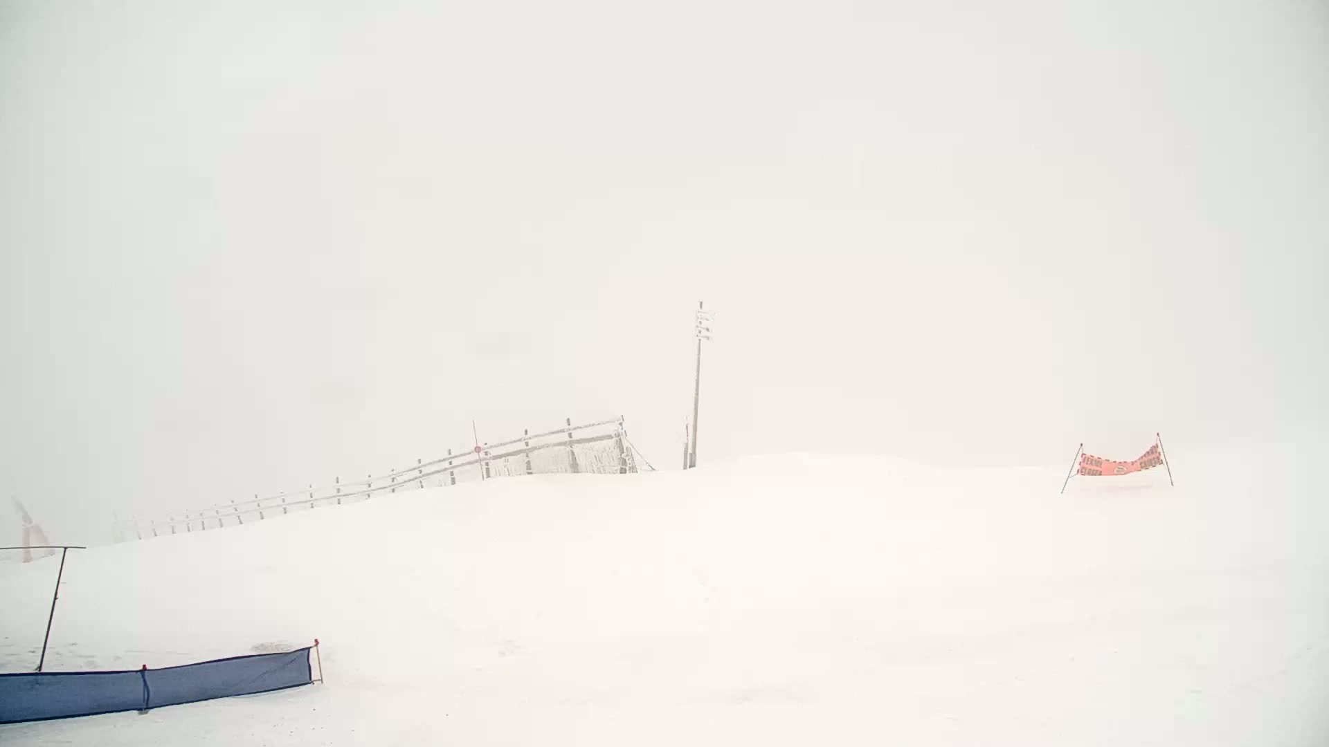 A snow-covered landscape with a partially buried wooden fence, a utility pole, and a low-hanging orange barrier is visible in a heavy fog.
