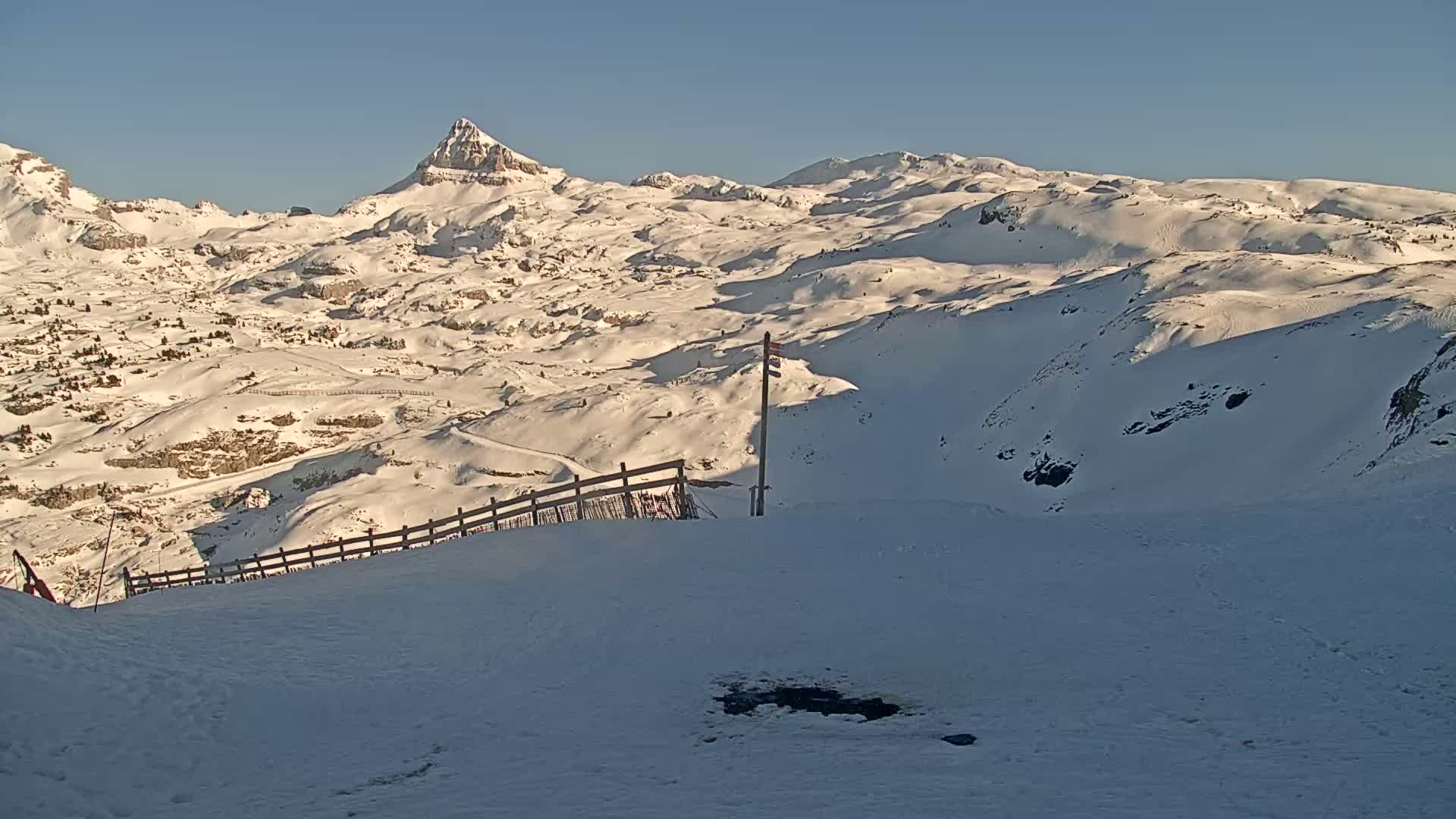 A snow-covered mountain range stretches out under a clear blue sky, with a wooden fence and a lone pole visible in the foreground.