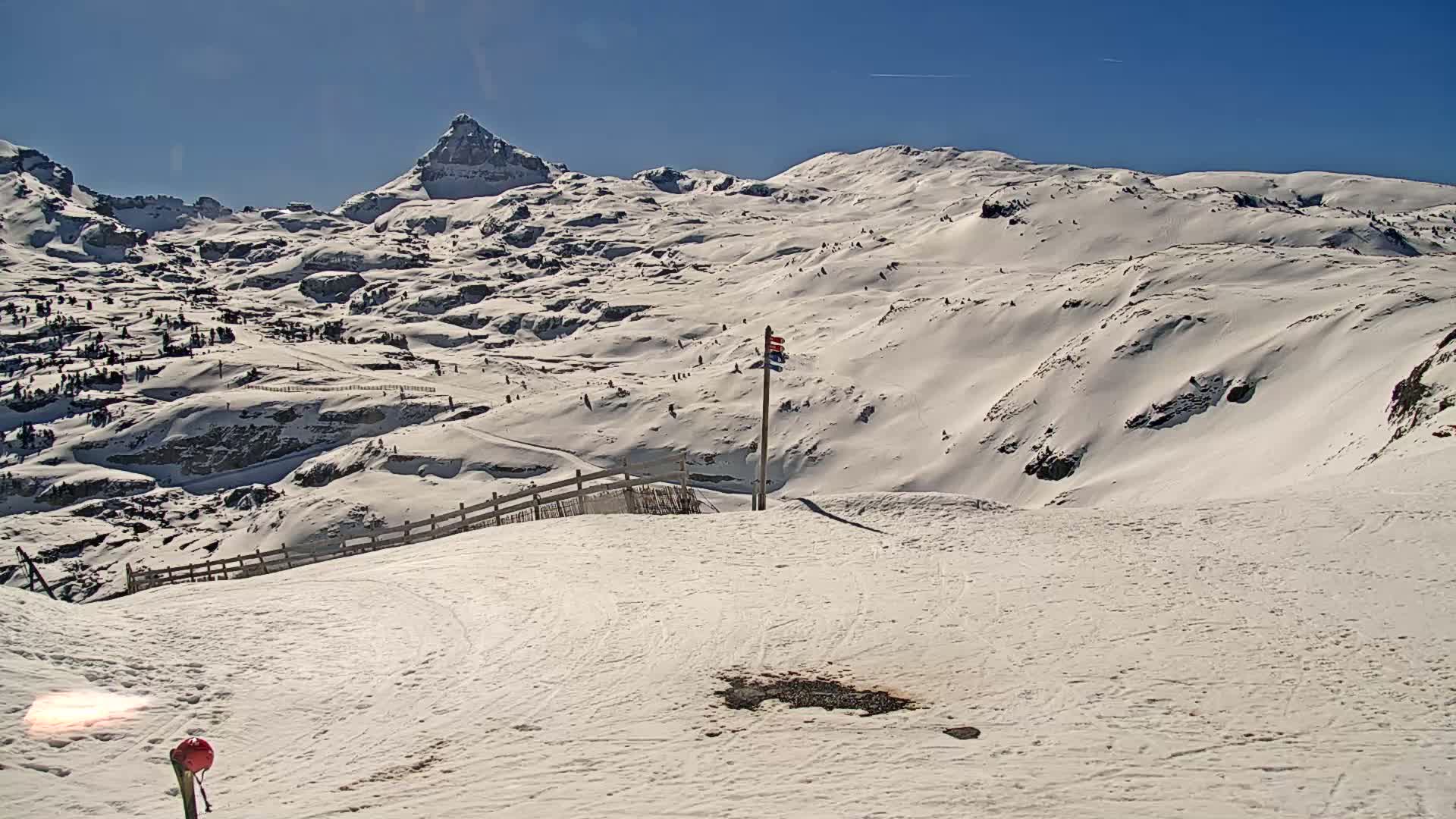 A snow-covered mountain range is visible under a bright, sunny sky, with a ski slope in the foreground featuring a small marker and fence.