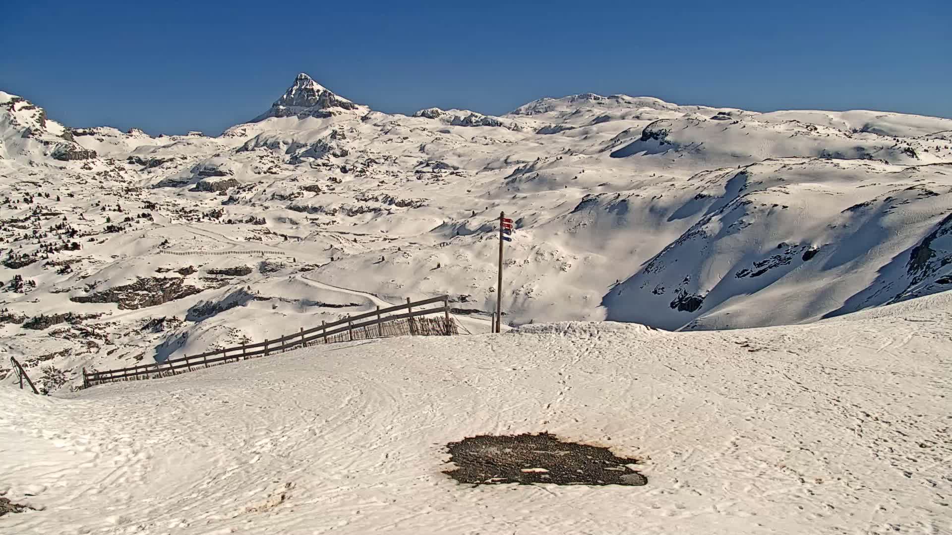 A snow-covered mountain landscape on a sunny day, featuring a prominent peak in the background and a wooden fence in the foreground.