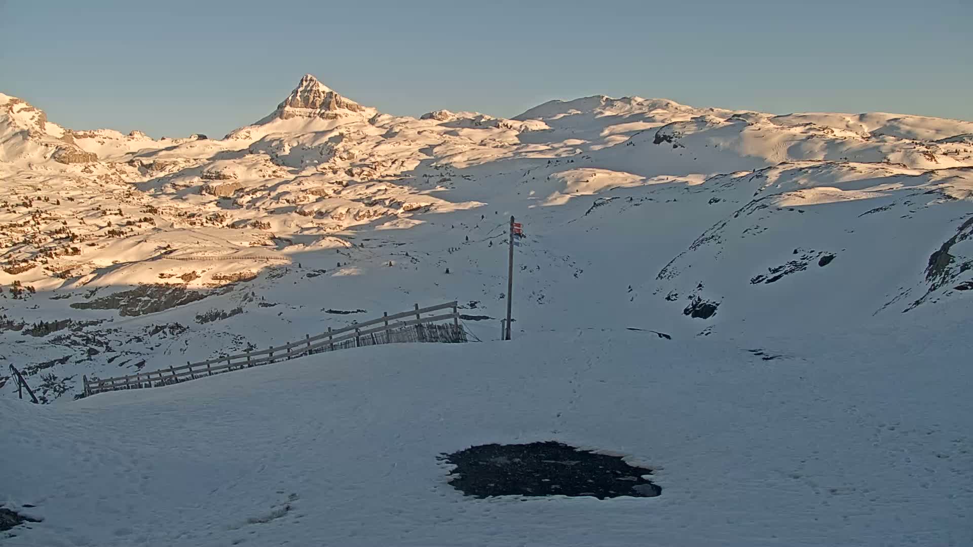 A snow-covered mountainous landscape under a clear, bright sky shows a small, dark patch of exposed ground near a wooden fence, with a distant peak prominently visible.