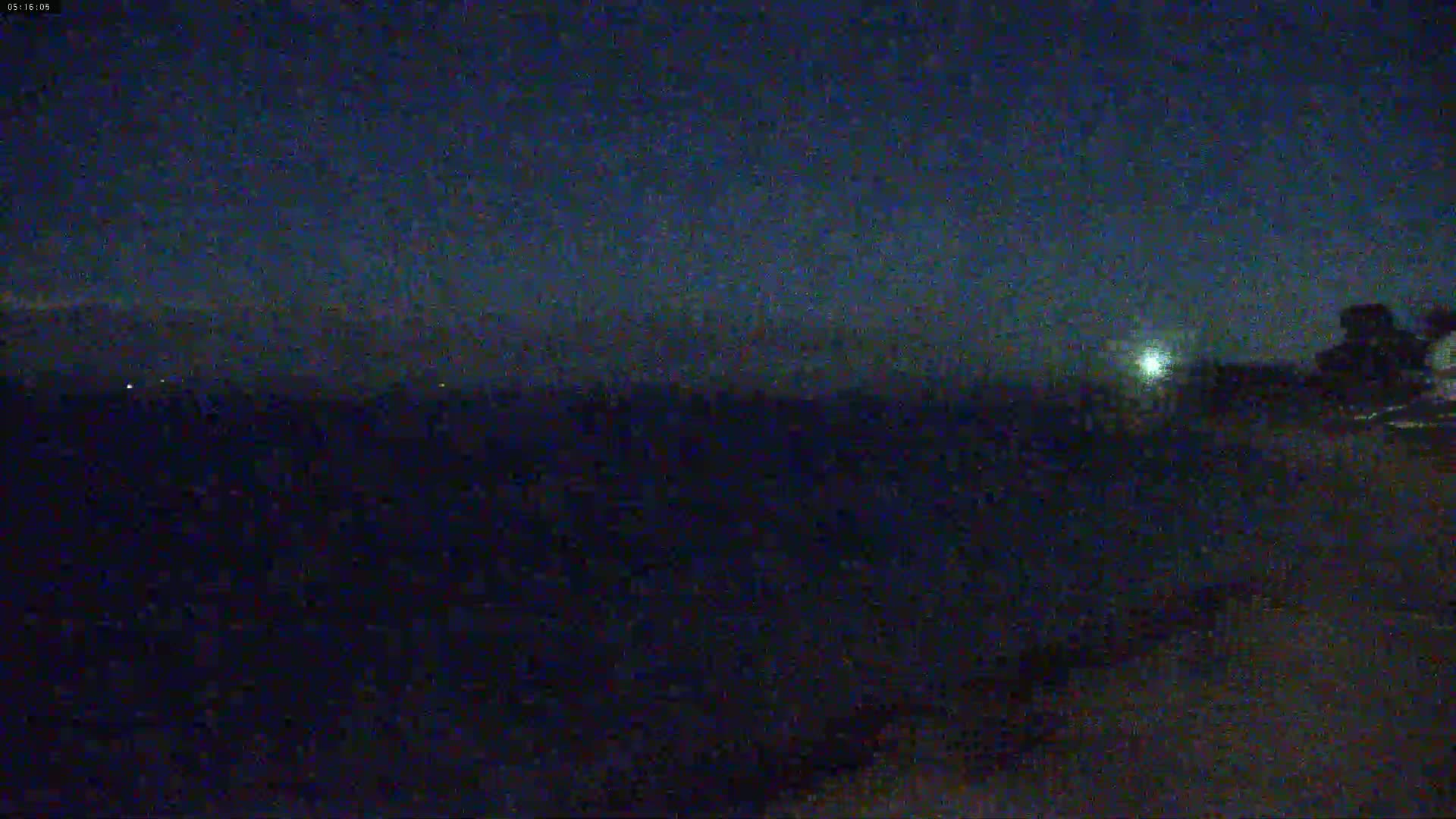 A dark, nighttime scene shows a dimly lit landscape under a mostly clear sky with a bright light source visible in the distance.