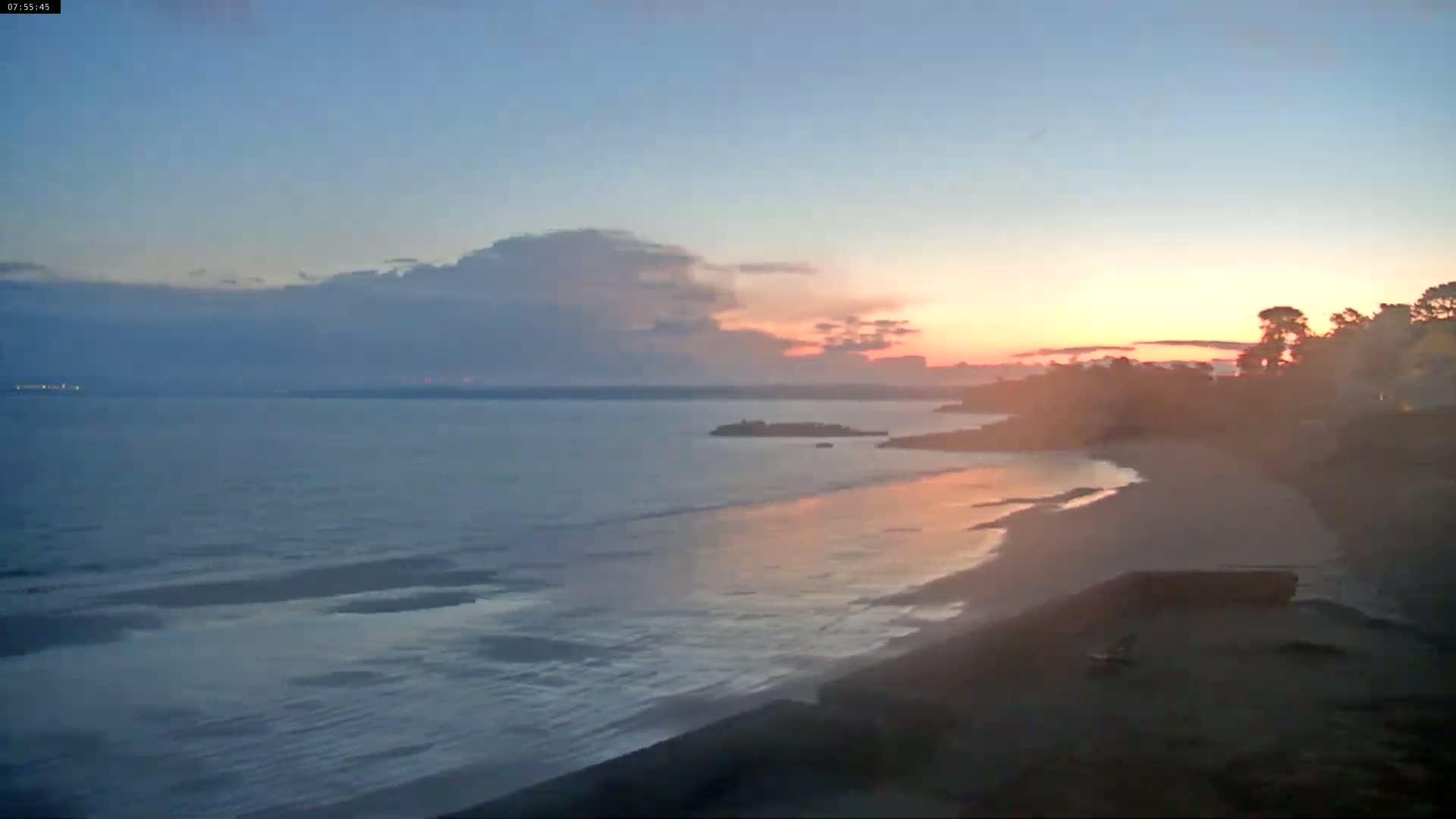 A calm ocean at sunset with a sandy beach and trees in the foreground under a partly cloudy sky.