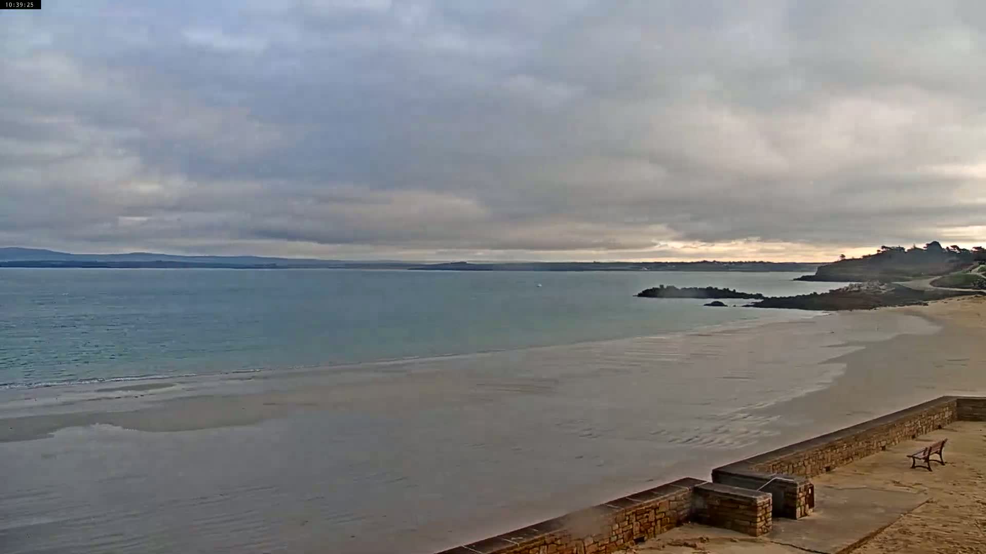 A wide, sandy beach stretches to a calm, light teal ocean under a cloudy sky.