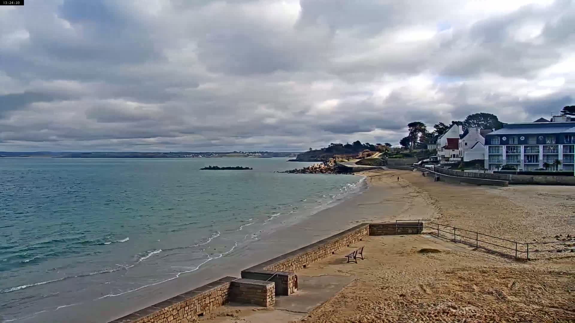 A sandy beach with gentle waves under a cloudy sky stretches toward buildings and a calm body of water.