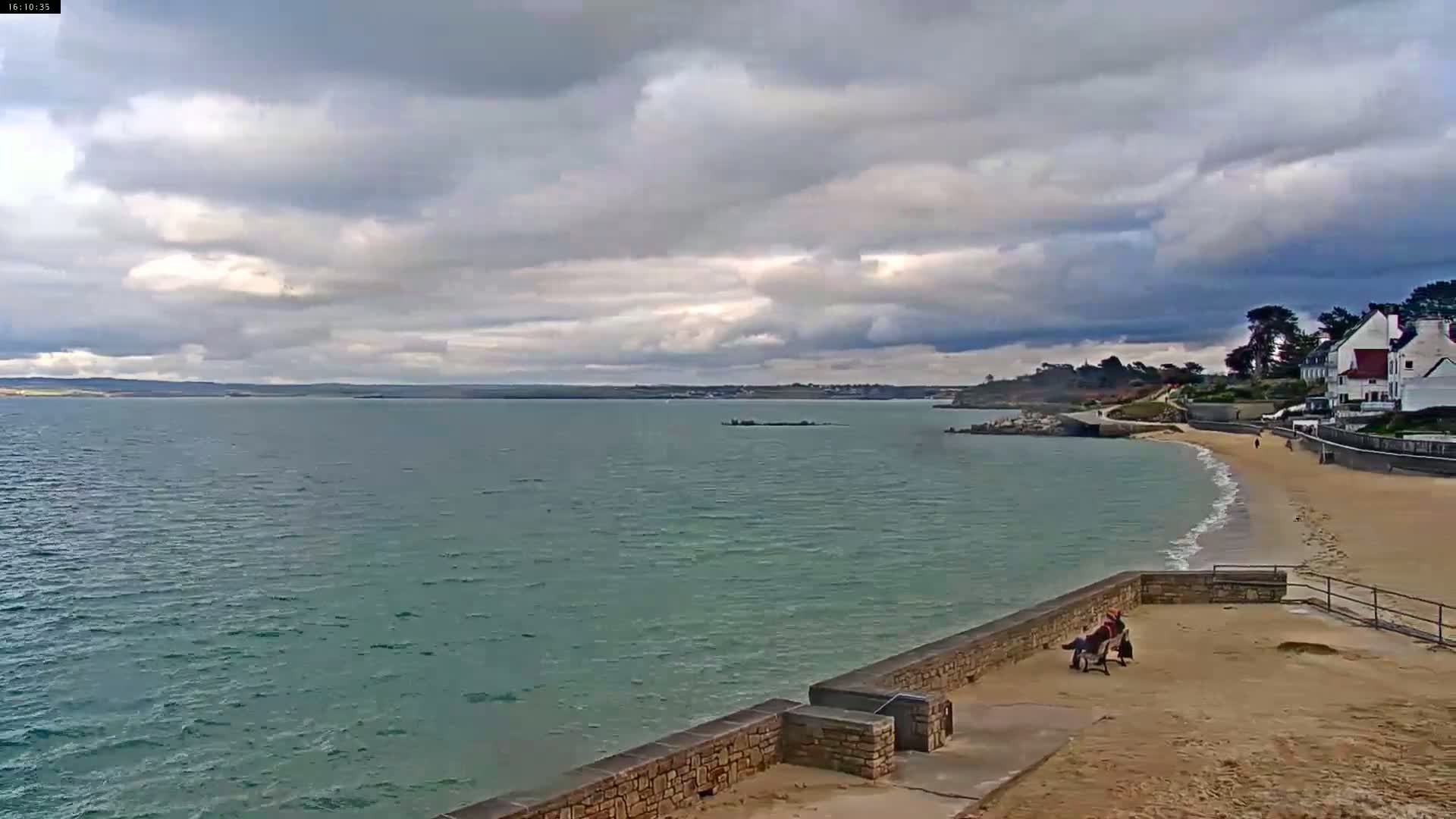A mostly cloudy day shows a calm sea next to a sandy beach with a stone wall and a few people sitting on a bench.