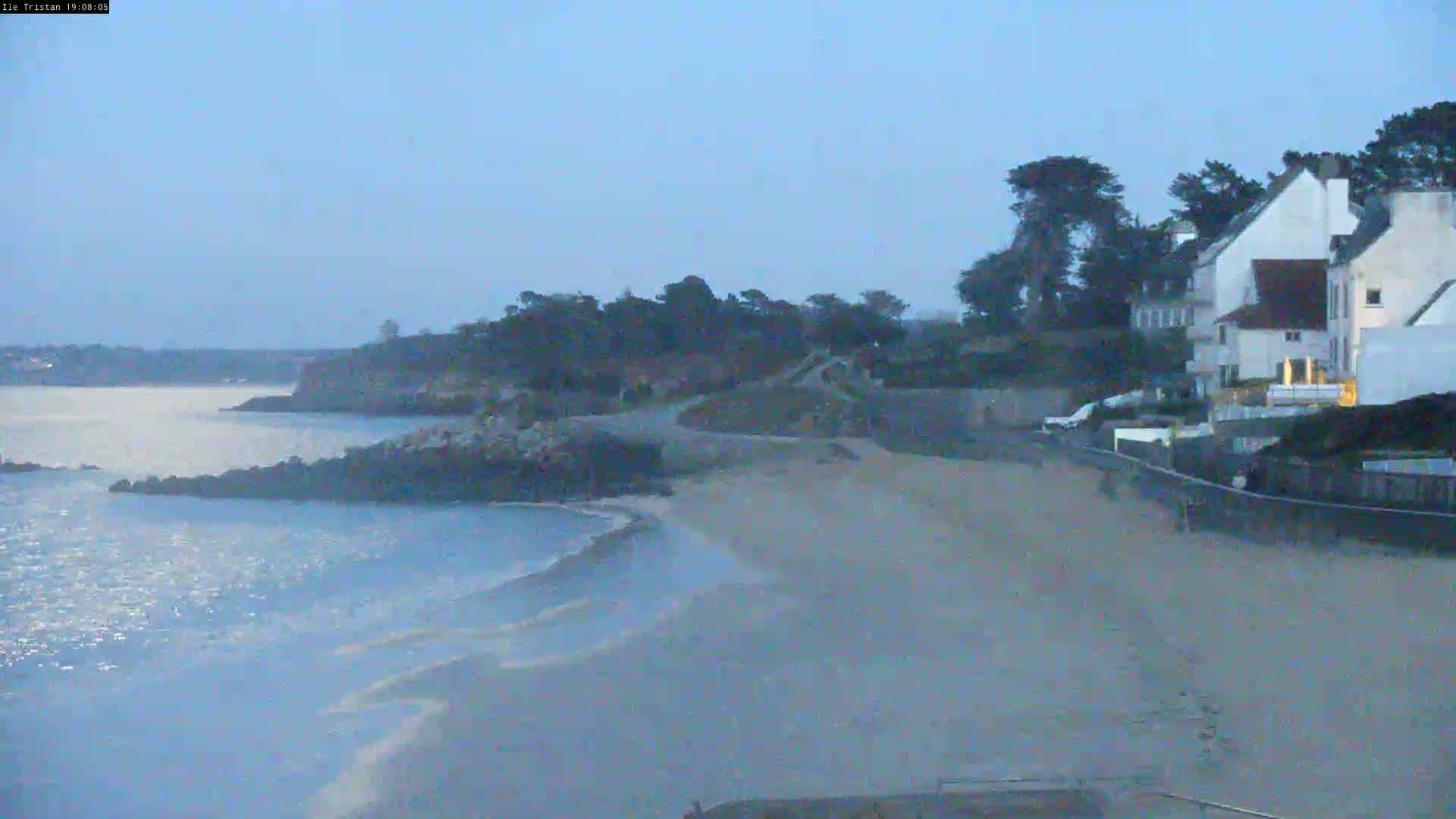 A sandy beach curves along a calm body of water under a clear, pale blue sky, with houses and trees visible on a hill overlooking the shore.