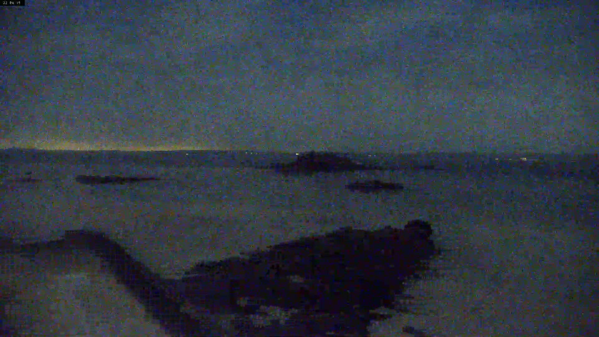 A dark, nighttime coastal scene shows a calm ocean with distant lights and rocky outcrops under a mostly clear, dusky sky.