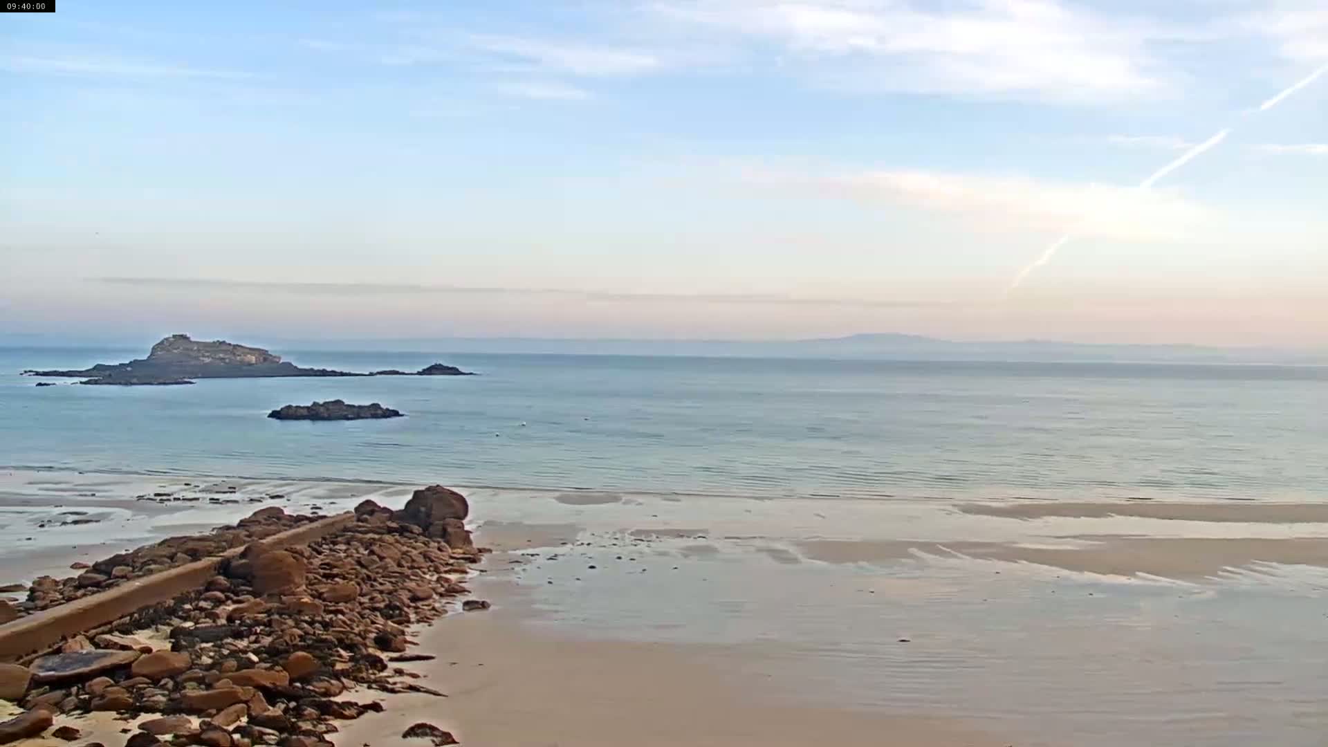 A sandy beach with calm, light-blue water and several rocky islands in the distance under a mostly clear, pale sky.