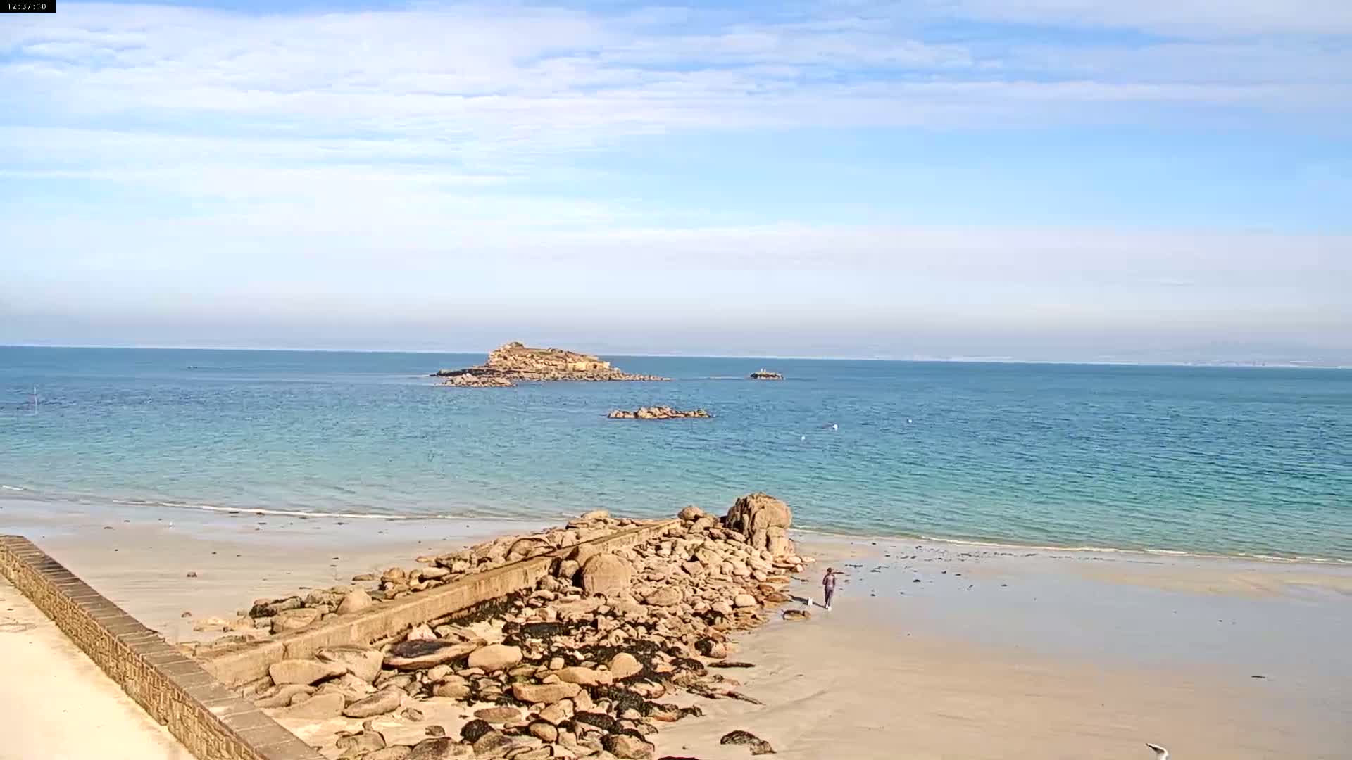A sandy beach with a rocky jetty extends into a calm, light-blue ocean under a mostly clear sky, with small islands visible in the distance.