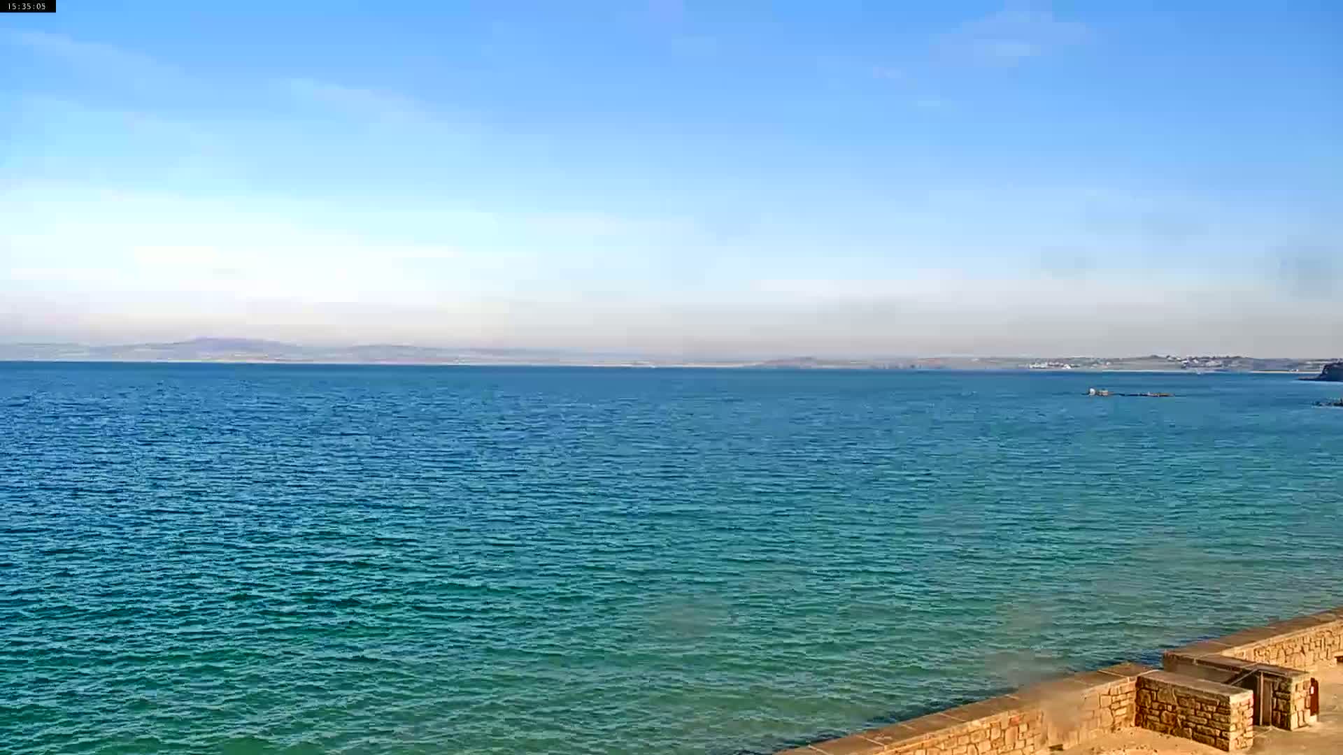 A tranquil teal sea stretches to a hazy horizon under a clear blue sky, with a low stone wall in the foreground.