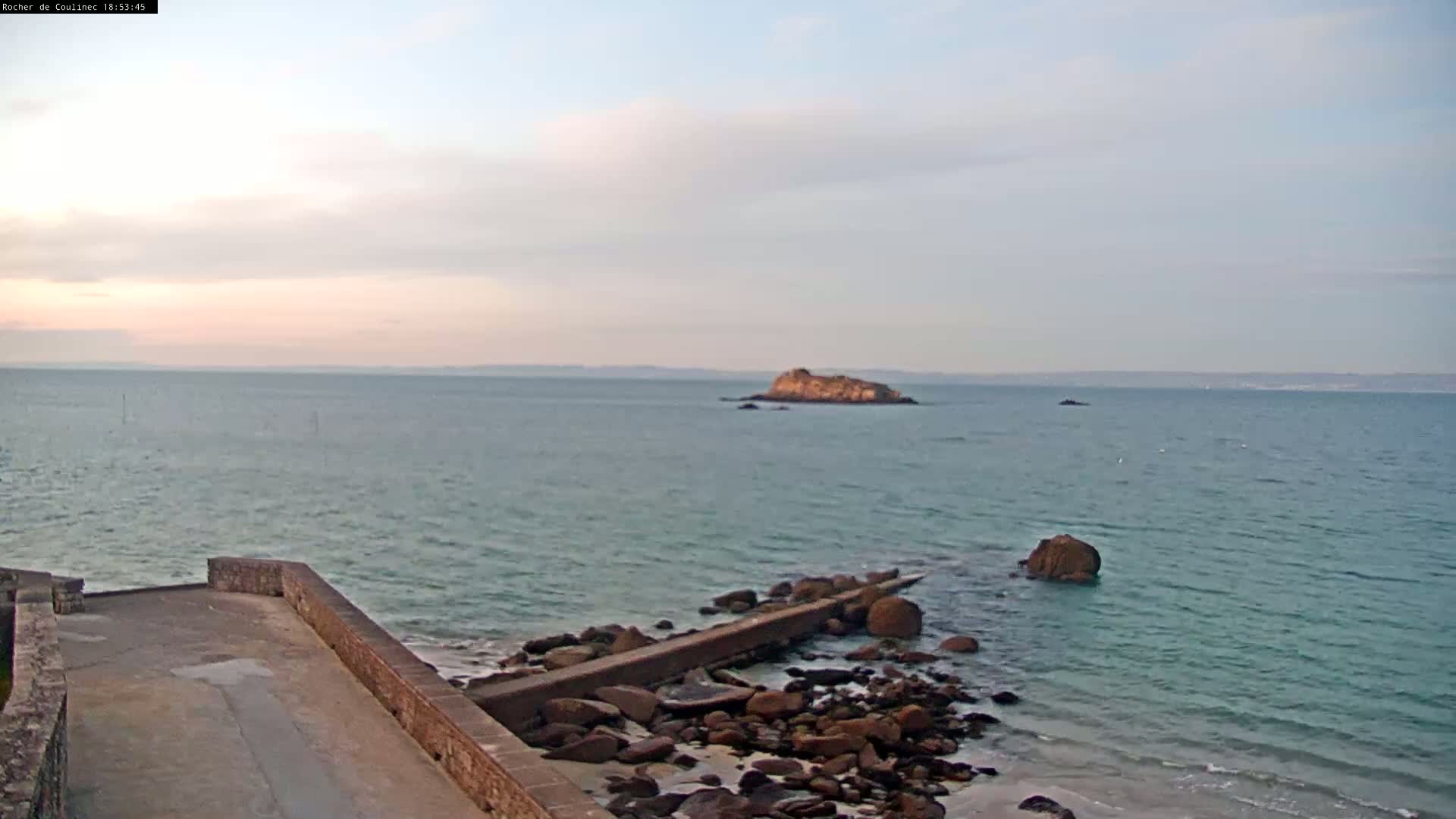 A rocky shoreline with a low concrete wall extends into a calm, light-blue sea toward a small island under a mostly cloudy sky.