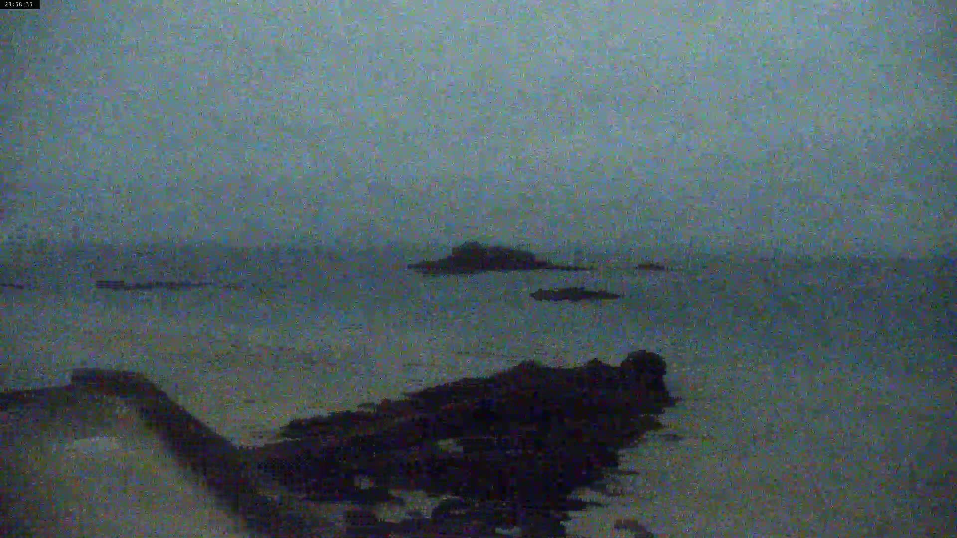 A dark, overcast sky hangs over a calm body of water where several dark rocks and a small island are visible near the shoreline.