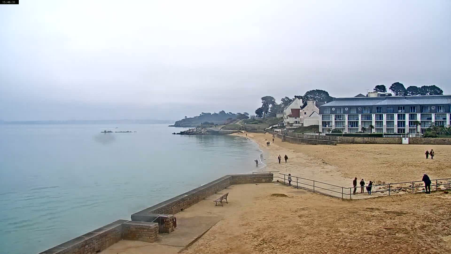 A sandy beach with several people walking on it and a large building behind it is visible, all under an overcast sky.