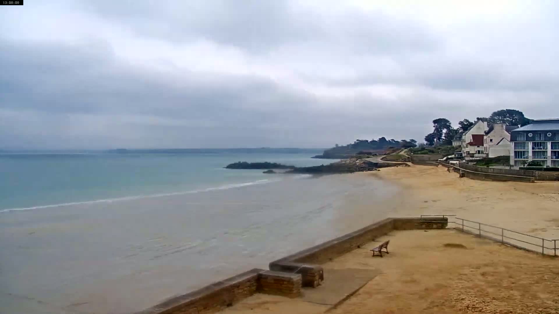 A sandy beach with calm, light-blue water stretches out under a cloudy sky, bordered by a low stone wall and buildings.