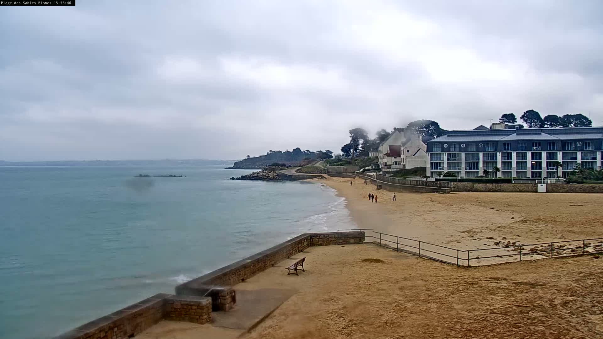 A sandy beach with a few people walking along the shoreline, backed by buildings and a calm sea under an overcast sky.