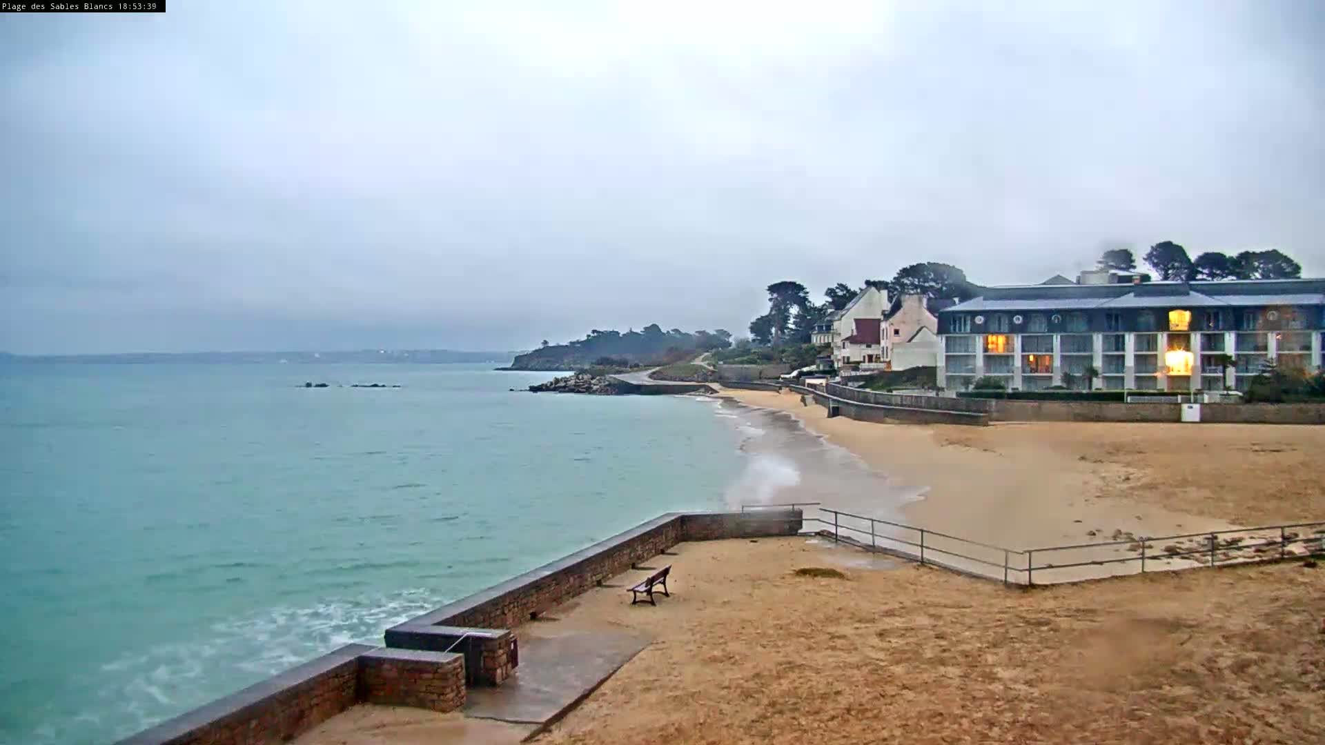 A sandy beach with a low brick wall and a hotel overlooks a calm, light-green sea under an overcast sky.