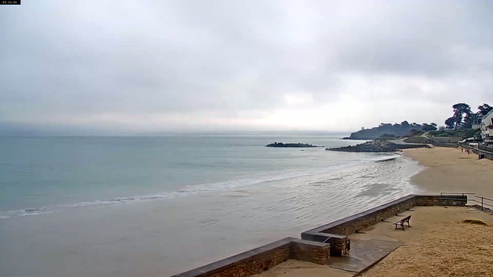 A sandy beach with gentle waves laps the shore under a cloudy sky, with a stone wall and buildings visible in the distance.