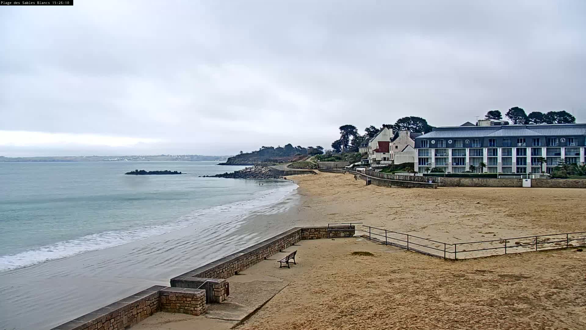 A sandy beach with a low stone wall and a few buildings behind it on an overcast day, with calm waves lapping the shore.