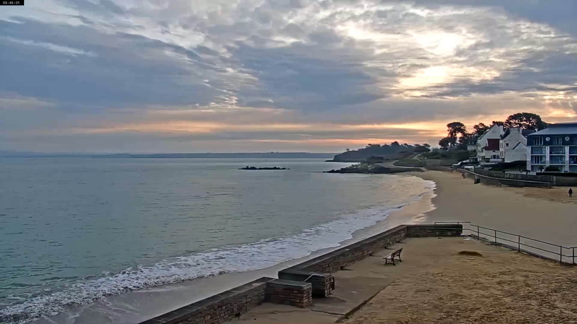 A sandy beach with calm waves laps gently against a low stone wall under a partly cloudy sky at sunrise.