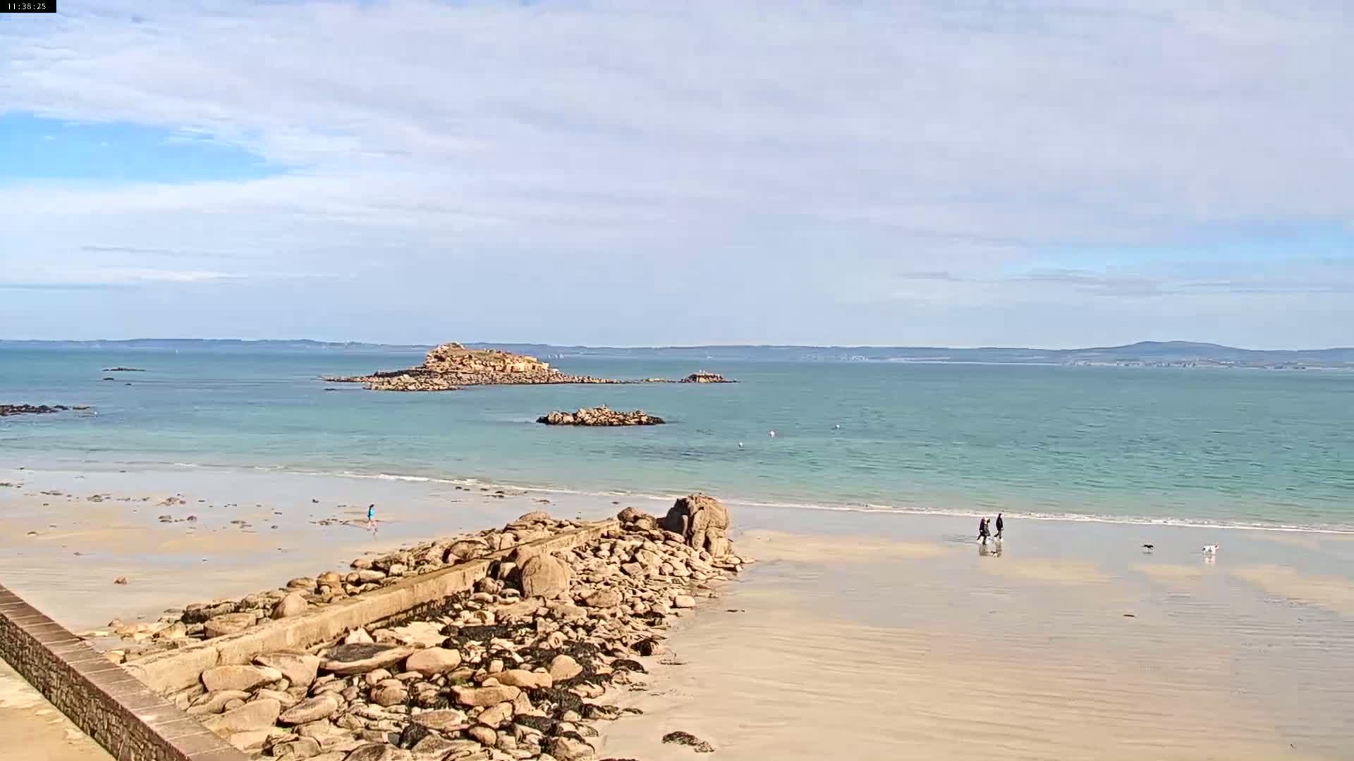 A sandy beach with a rocky jetty stretches towards a calm, light-blue ocean with a small island in the distance under a partly cloudy sky.