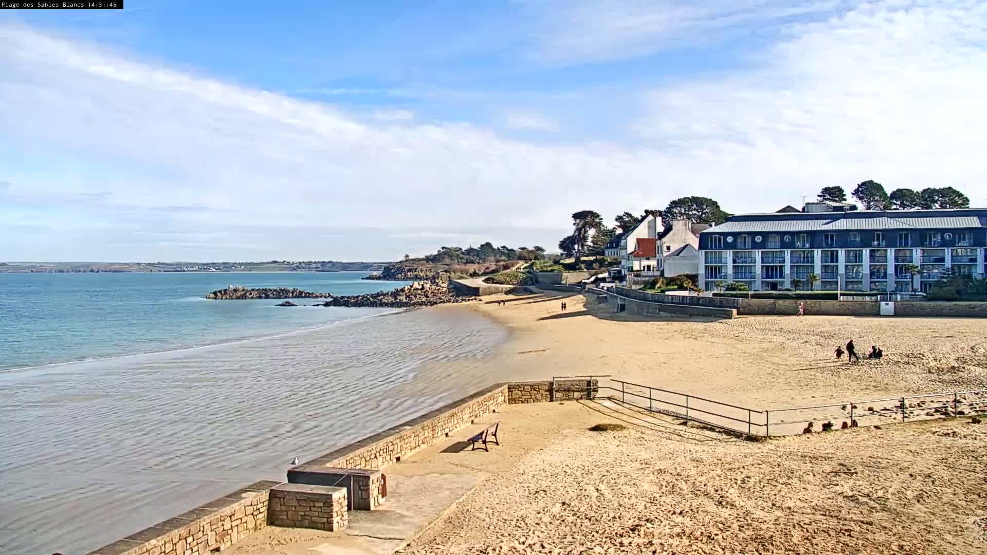 A sandy beach with calm, light blue water stretches out to a calm sea under a mostly sunny sky, next to a multi-story building and some smaller homes.