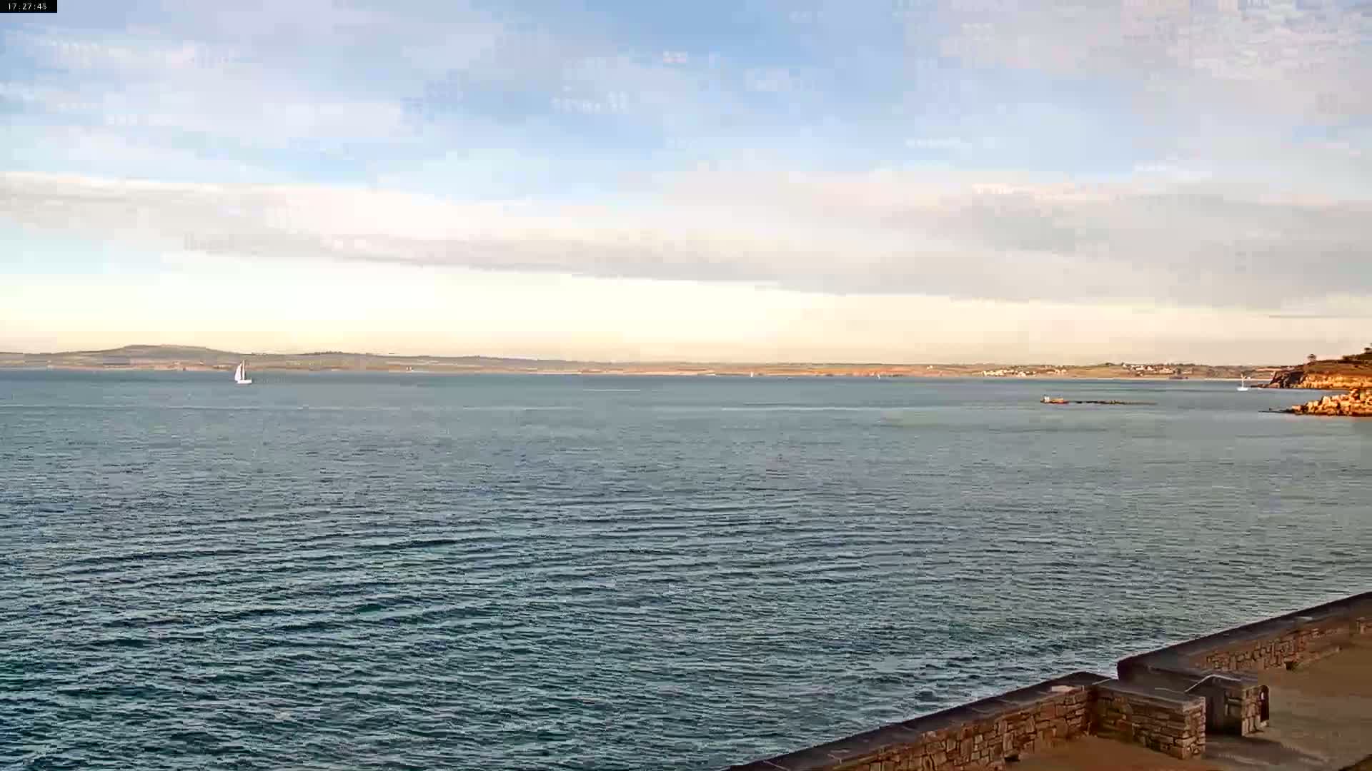 A calm sea with a few small boats is viewed from a stone wall on a partly cloudy day.