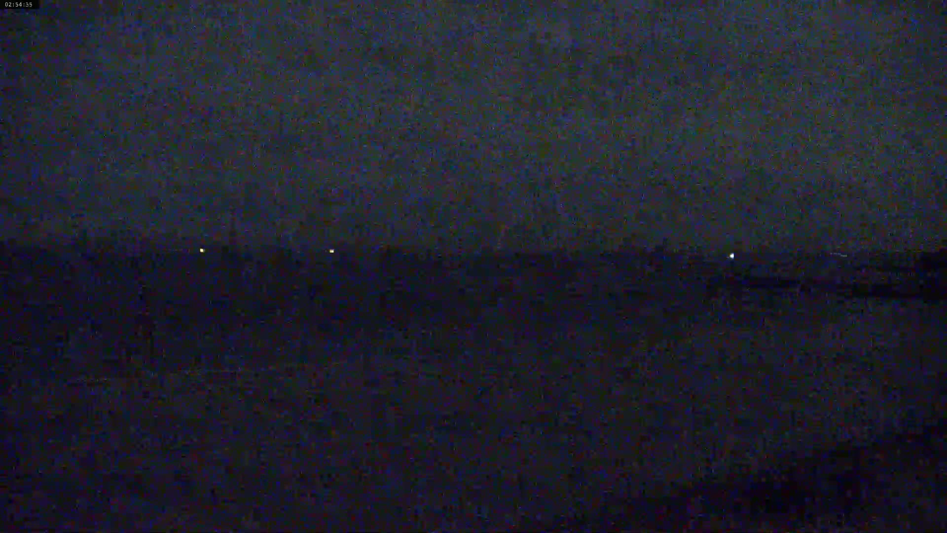A dark, nighttime scene shows a flat, dark landscape with a few distant, small lights under a mostly clear, dark sky.