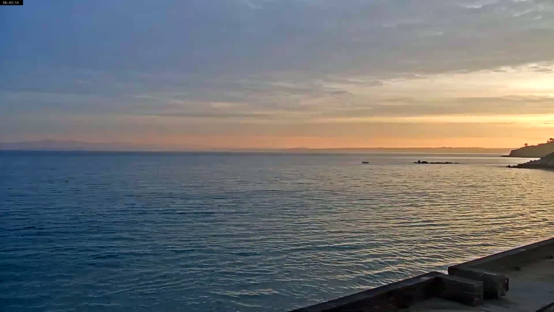 A calm ocean at sunset is viewed from a coastal vantage point, with a distant landmass visible on the horizon.