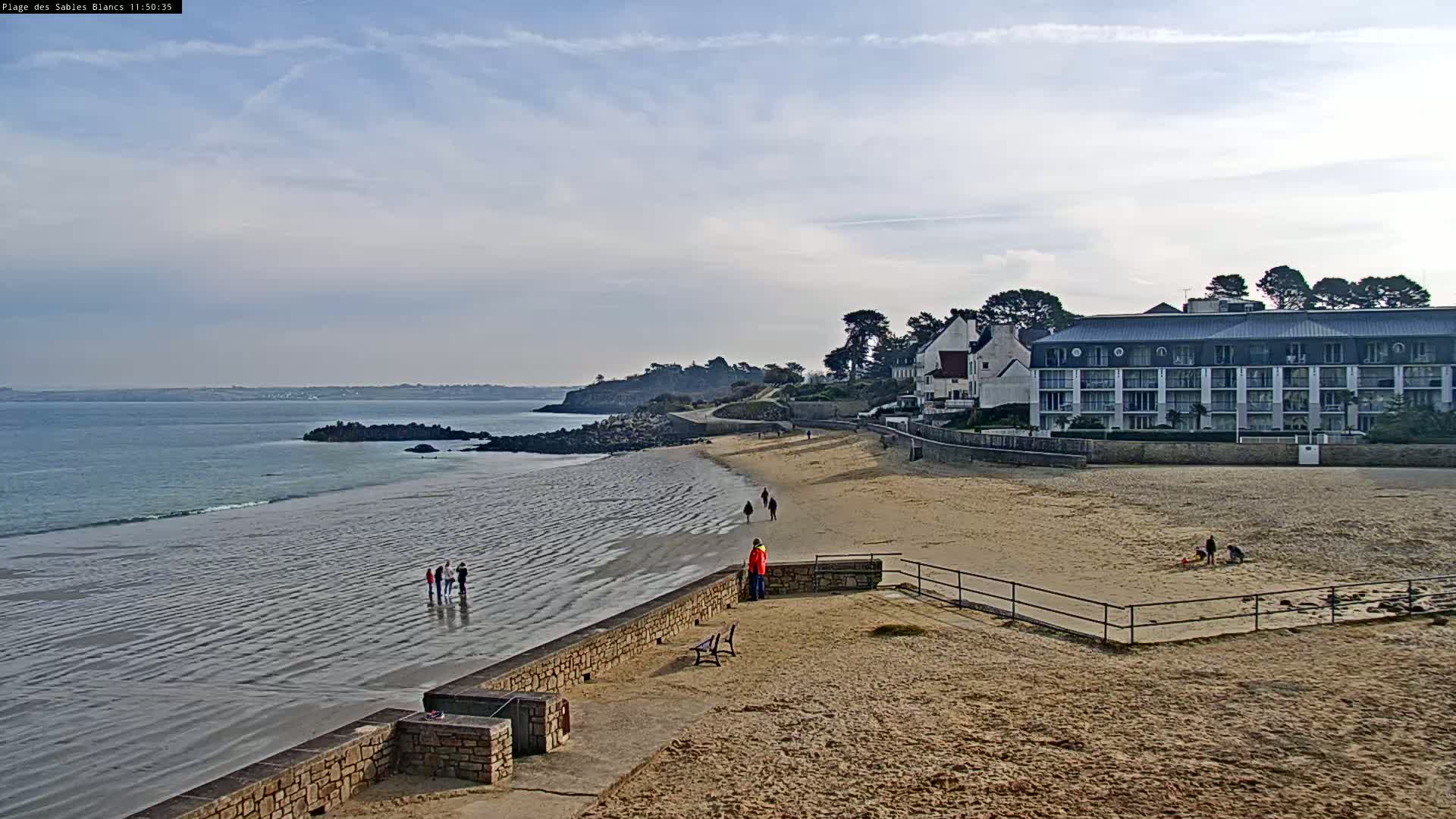 A sandy beach with a few people walking on it, next to a calm ocean under a partly cloudy sky, and a multi-story building in the background.