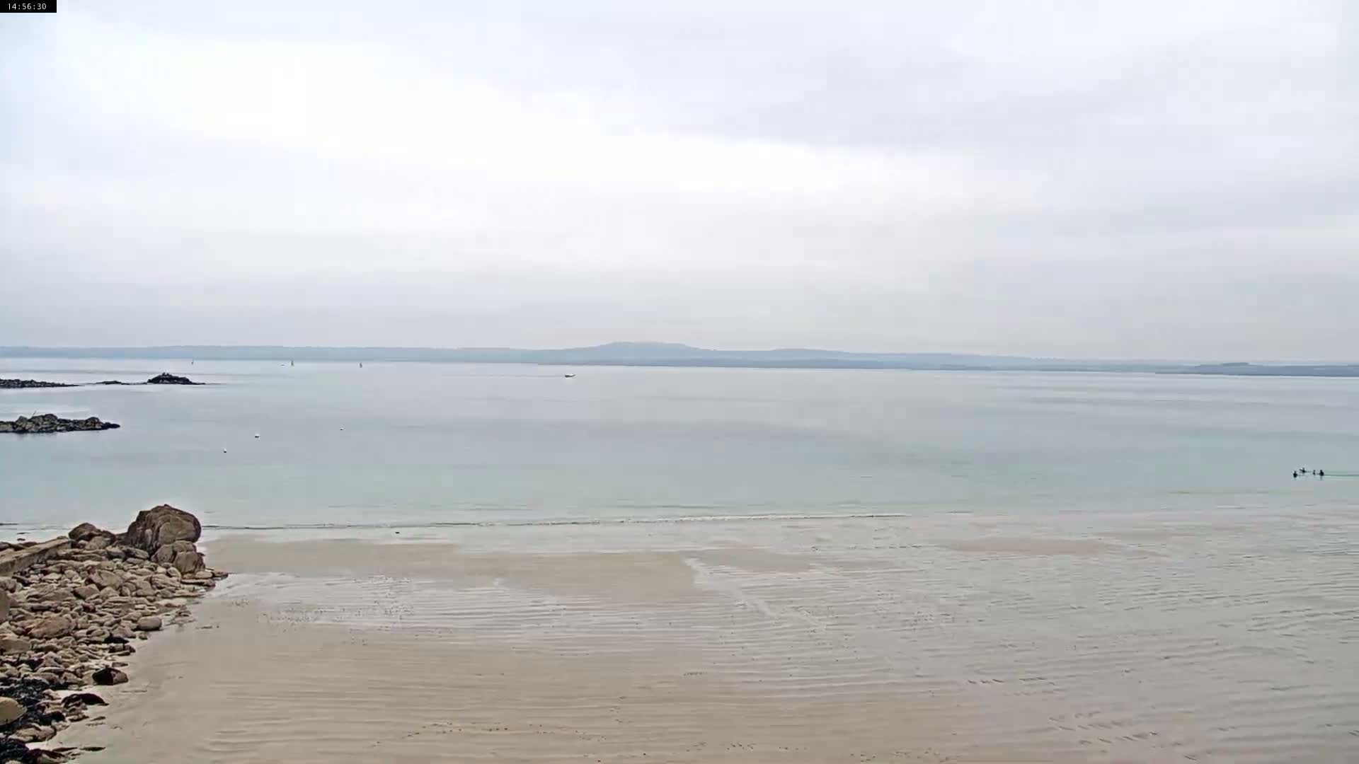 A sandy beach slopes gently into calm, light-blue water under an overcast sky, with a distant shoreline visible.
