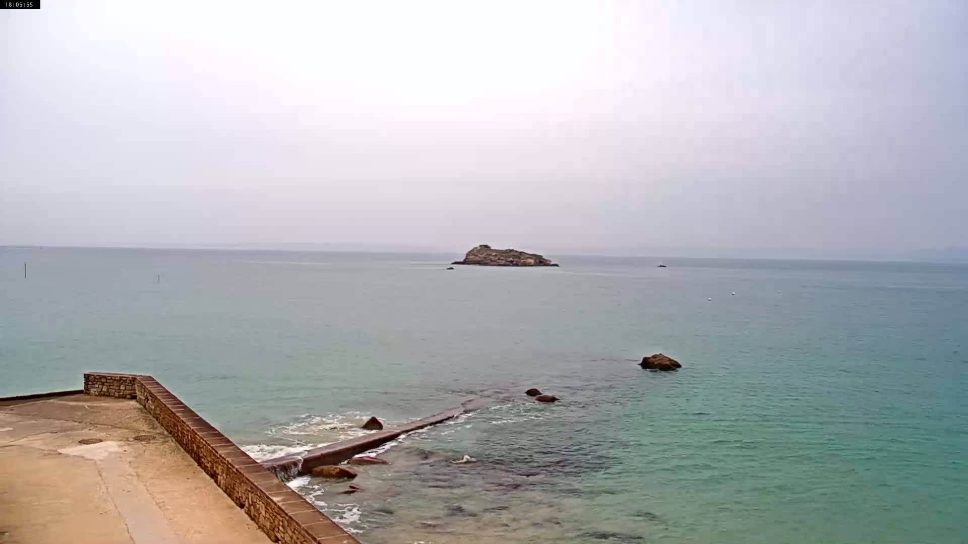 A rocky islet sits in a calm, light-green sea under an overcast sky, viewed from a stone seawall.