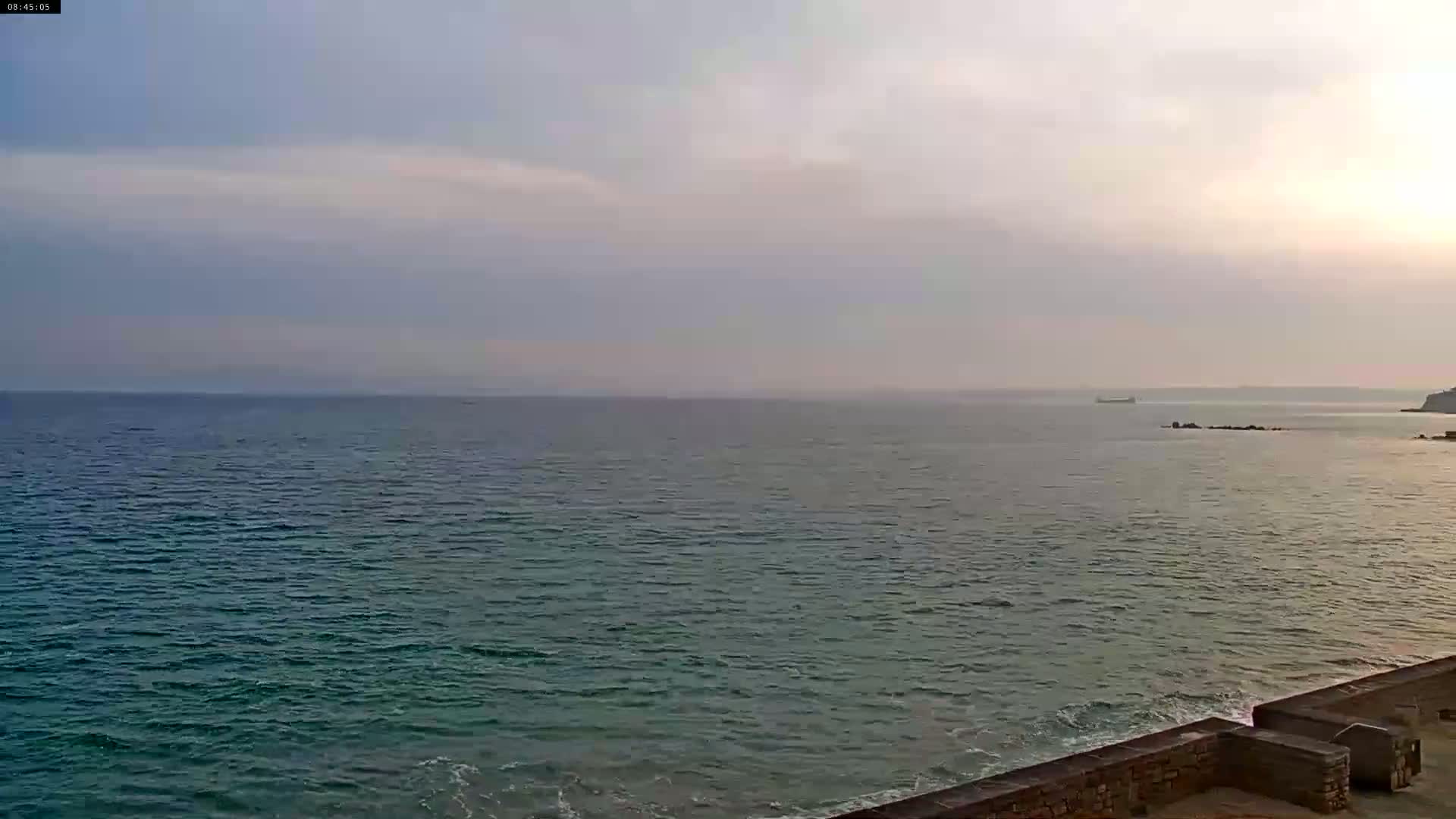 A calm sea under a hazy, light-colored sky meets a low stone wall in the foreground.