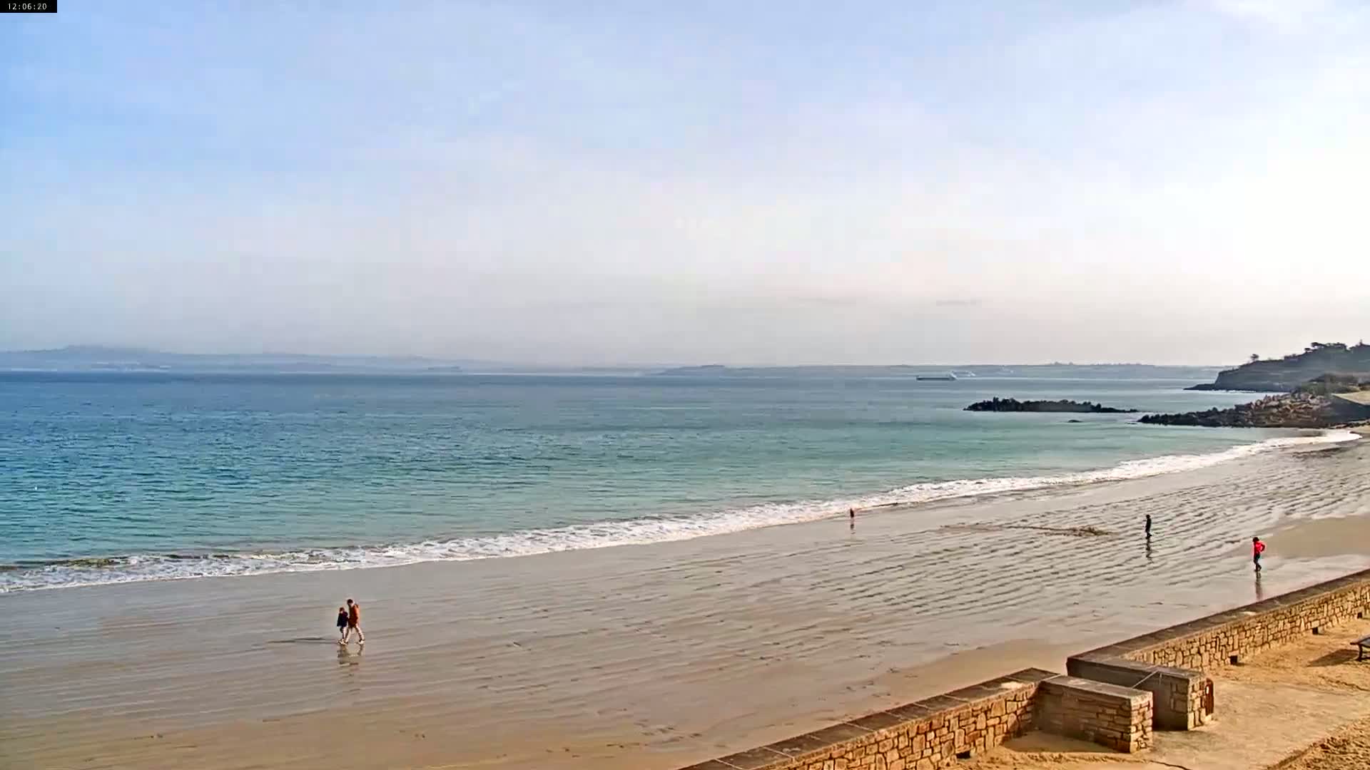 A wide sandy beach with gentle waves and a few people walking along the shore on a clear, sunny day, with calm waters and distant land visible on the horizon.