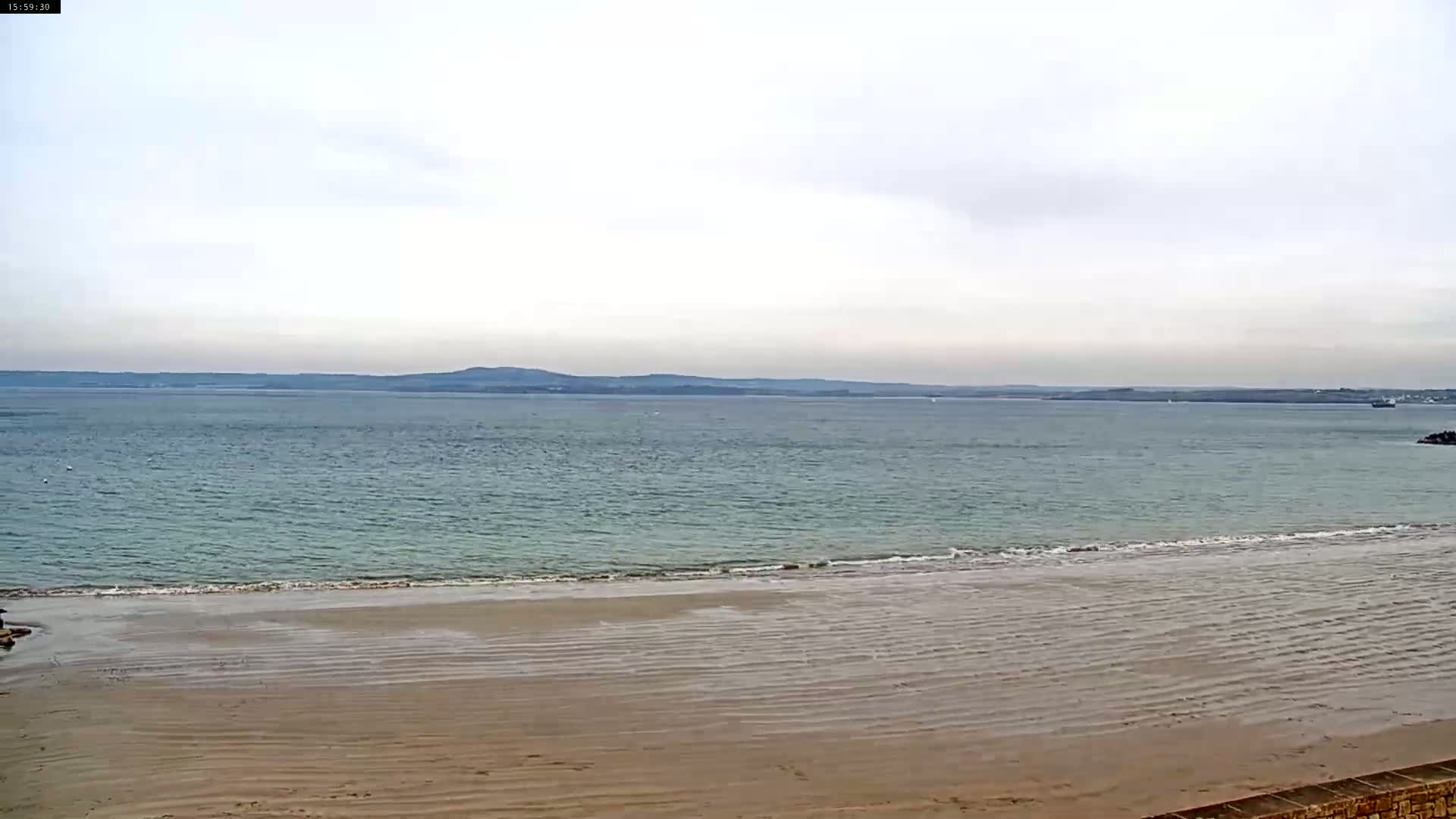 A sandy beach stretches to a calm, light-blue ocean under a mostly cloudy sky, with a distant shoreline visible on the horizon.