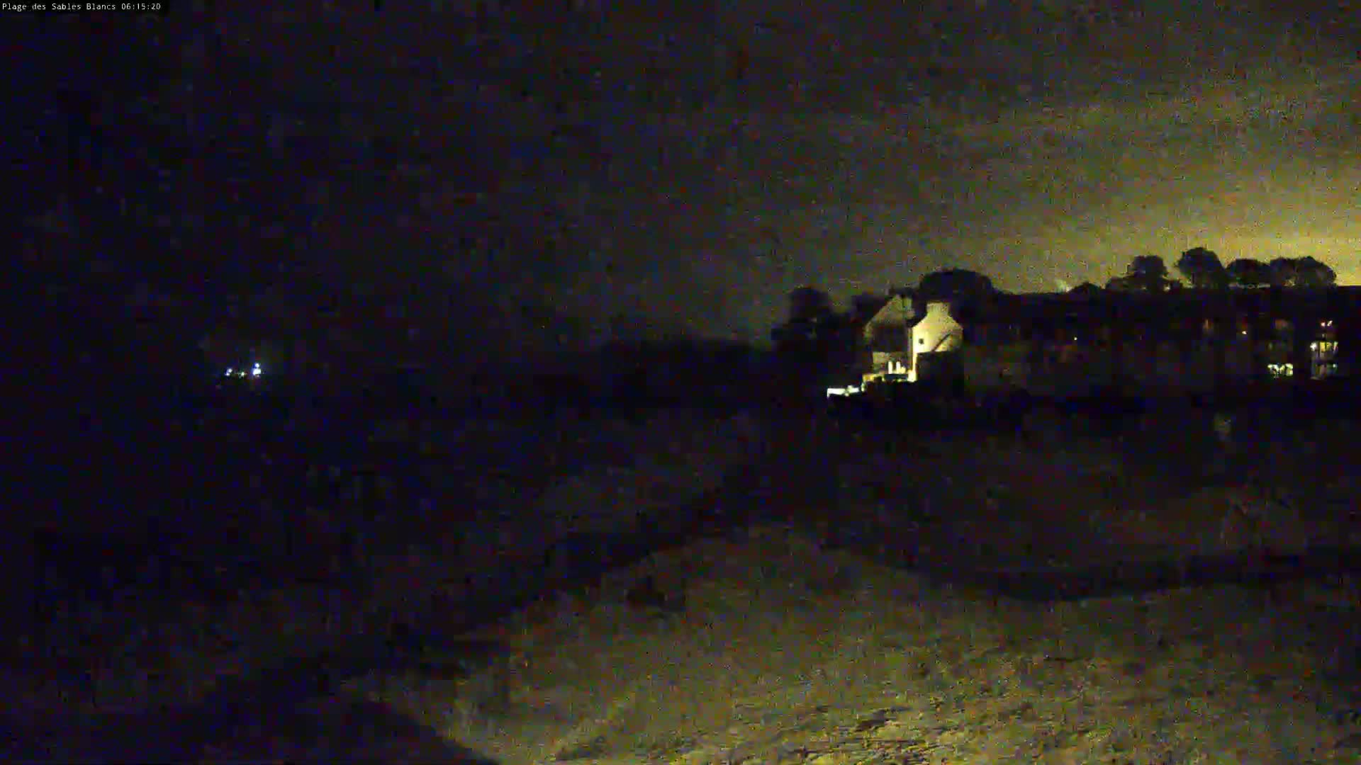At night, under a dark, cloudy sky, dimly lit buildings are visible in the distance, with a darker foreground that appears to be ground.